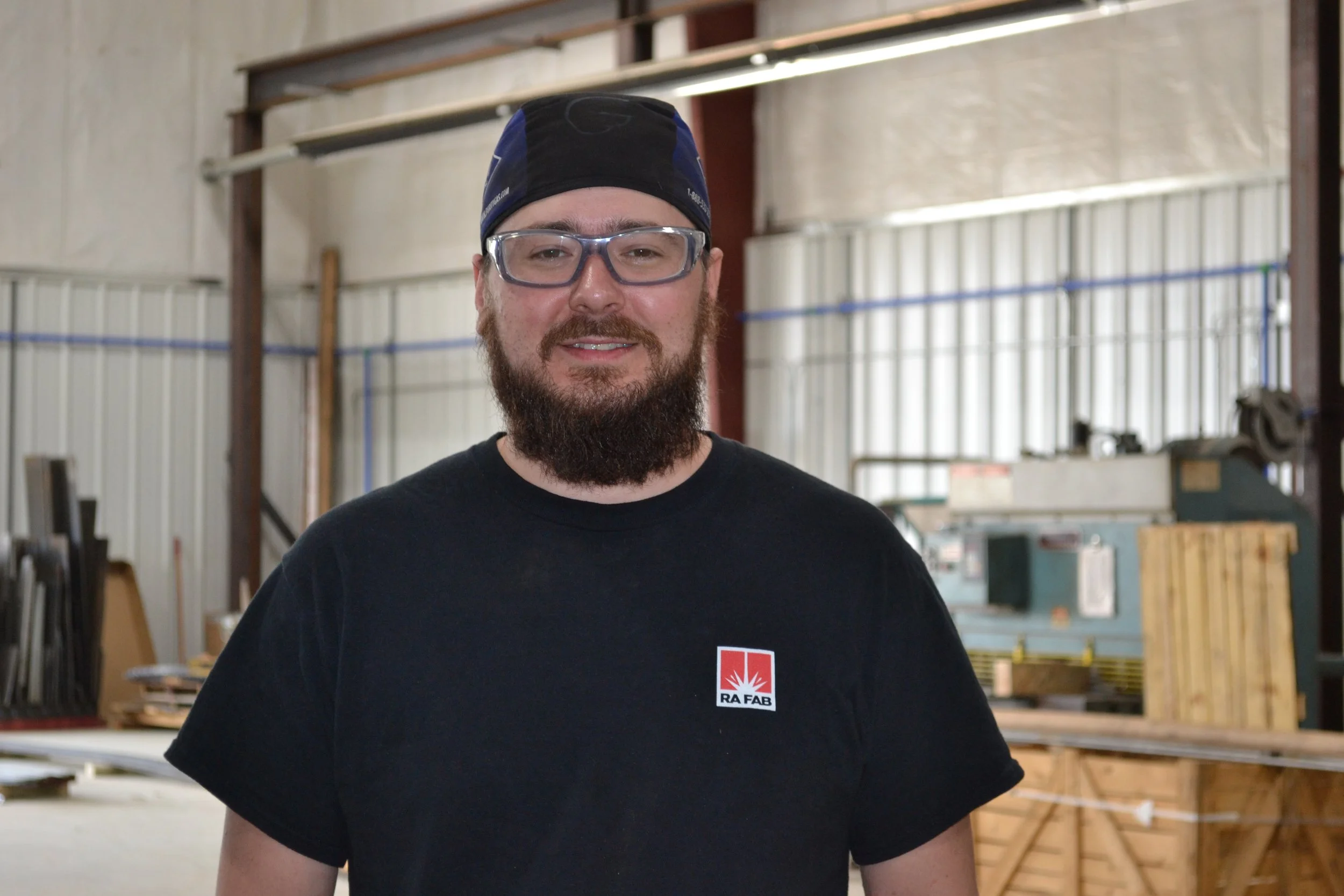 A man working on a metal frame structure in a workshop, wearing safety glasses and a bandana, with tools and papers on the work table.