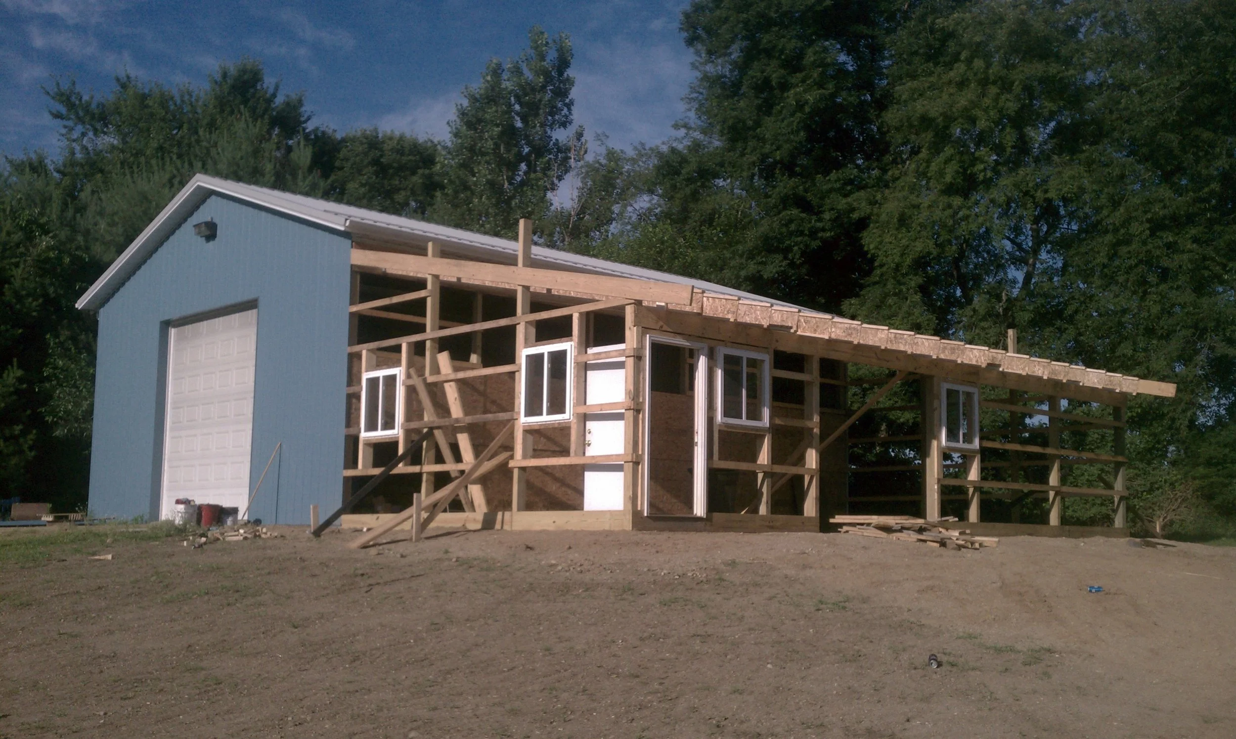 A pool barn under construction with a blue siding section on the left and an unfinished wooden structure on the right, featuring open window frames and door openings.