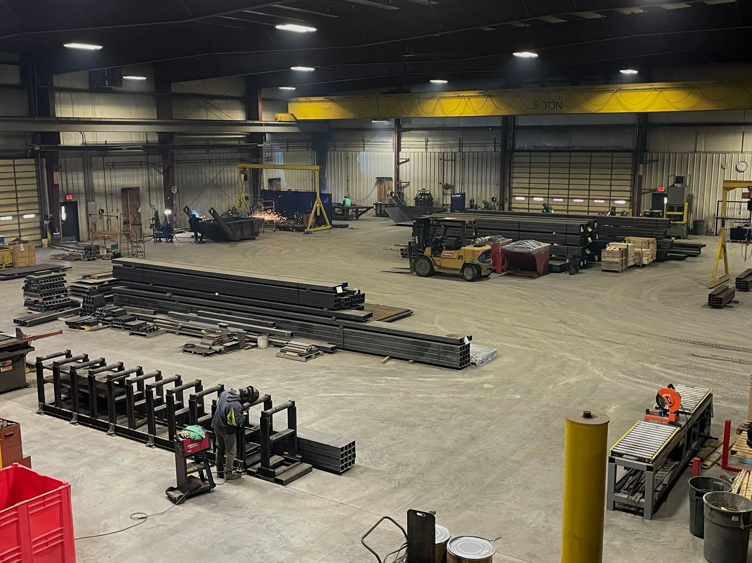 Interior of a large industrial warehouse with workers welding, stacks of metal pipes, and various construction materials.