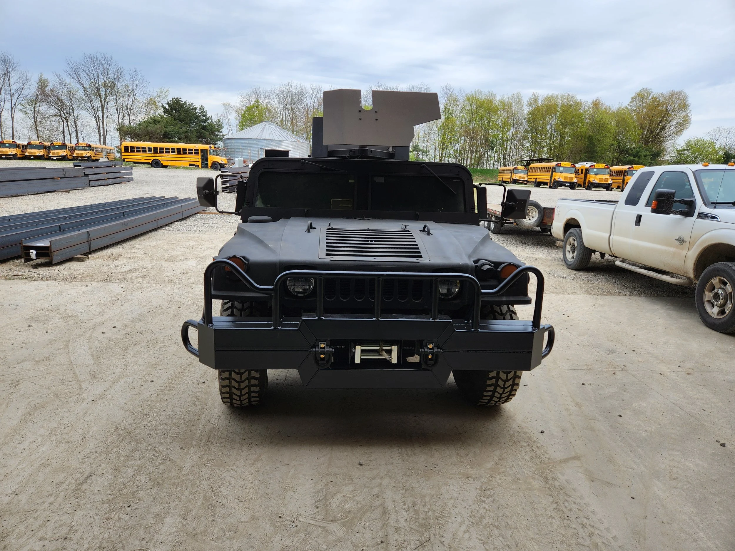 Black military-style vehicle with armored exterior and front bumper guard, parked on dirt ground, surrounded by trucks and school buses in a lot.
