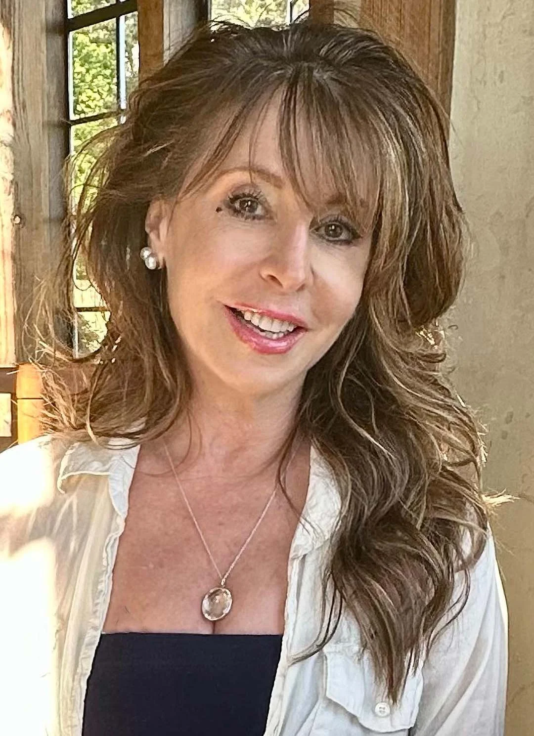 A woman with long, wavy brown hair and light skin, smiling, wearing pearl earrings, a necklace with a large round pendant, a black top, and a white shirt, sitting near a window with greenery outside.