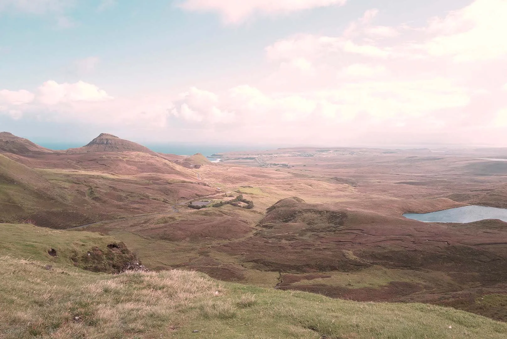Landscape with rolling hills, a small lake on the right, and a mountain in the background under a partly cloudy sky.
