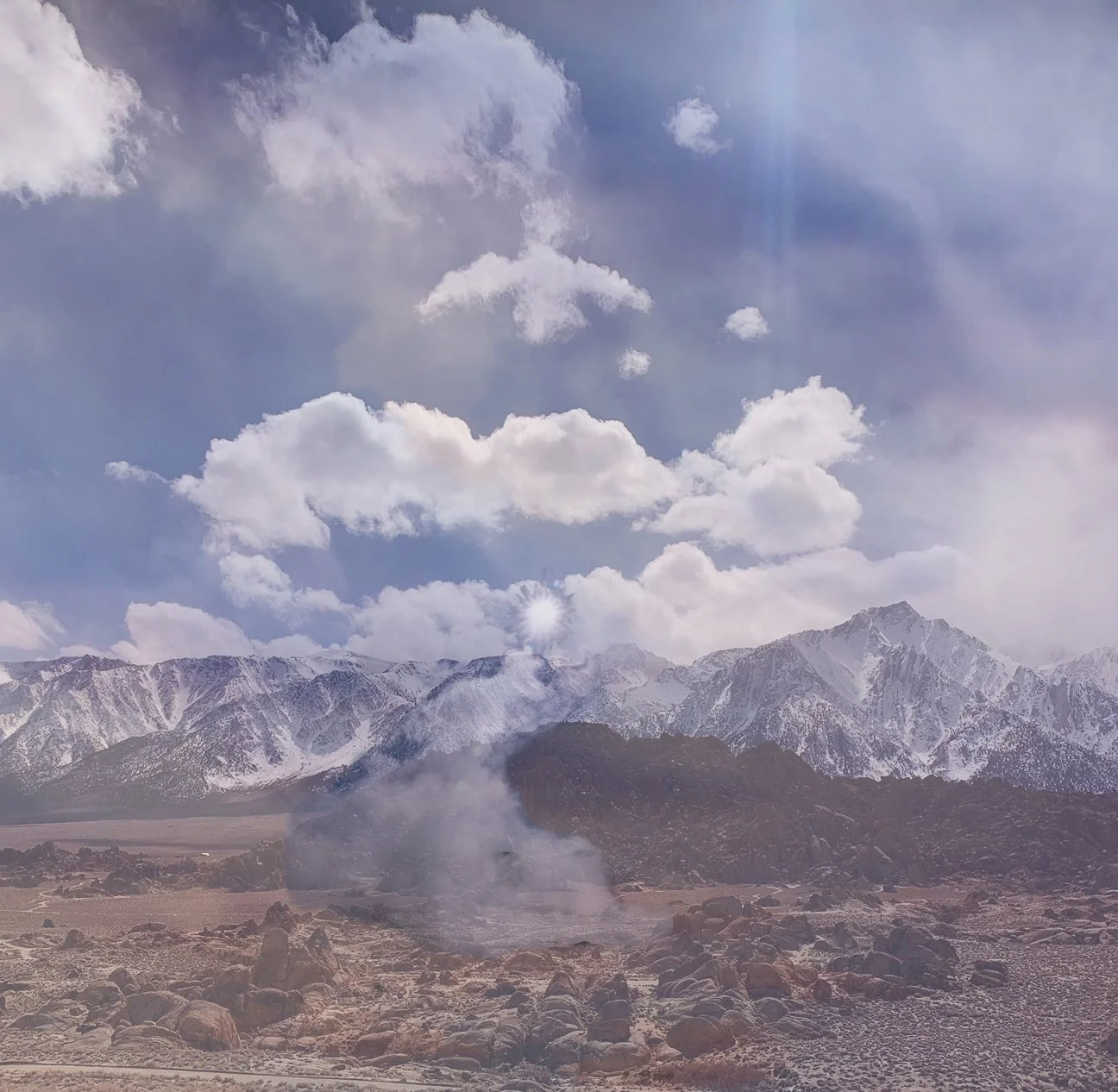 Snow-capped mountains under a partly cloudy sky with the sun shining through clouds over a rocky, arid plain with steam rising from the ground.