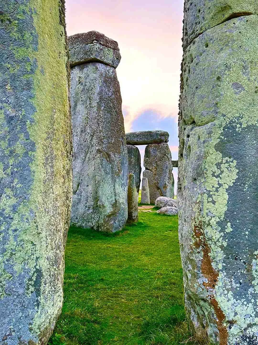 Stonehenge standing stones arranged in a row at sunrise or sunset, with moss and lichen on the stones and grass on the ground.