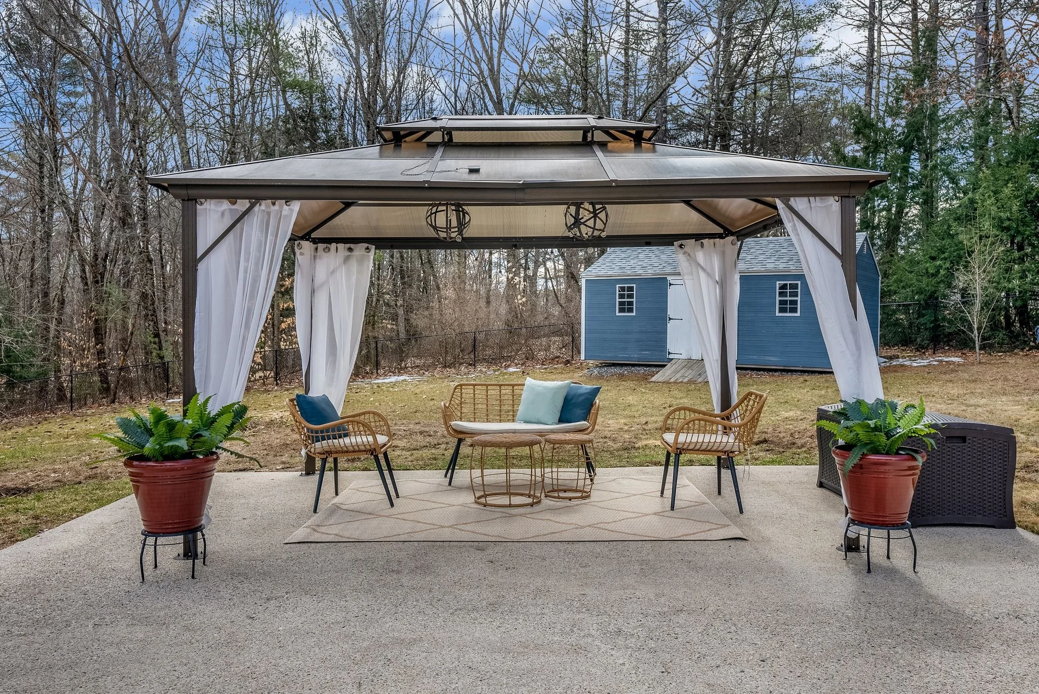 Outdoor gazebo with metal frame and white curtains, featuring wicker furniture with cushions, a rug on the concrete patio, and potted plants. Blue wooden shed and trees in the background.