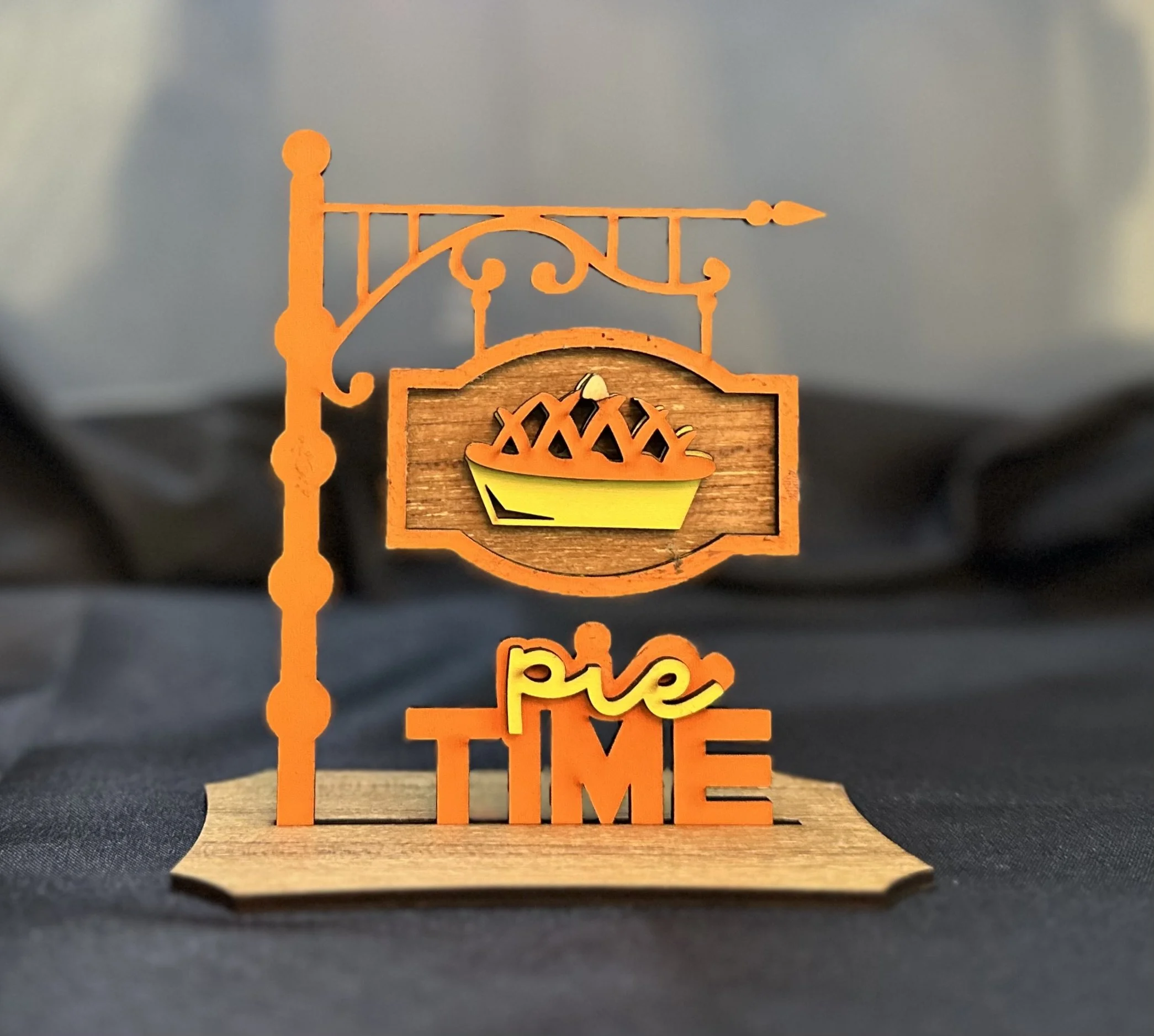 Decorative cake sign with a pie illustration, the word 'pie' in yellow cursive and 'TIME' in orange block letters, on a wooden base.