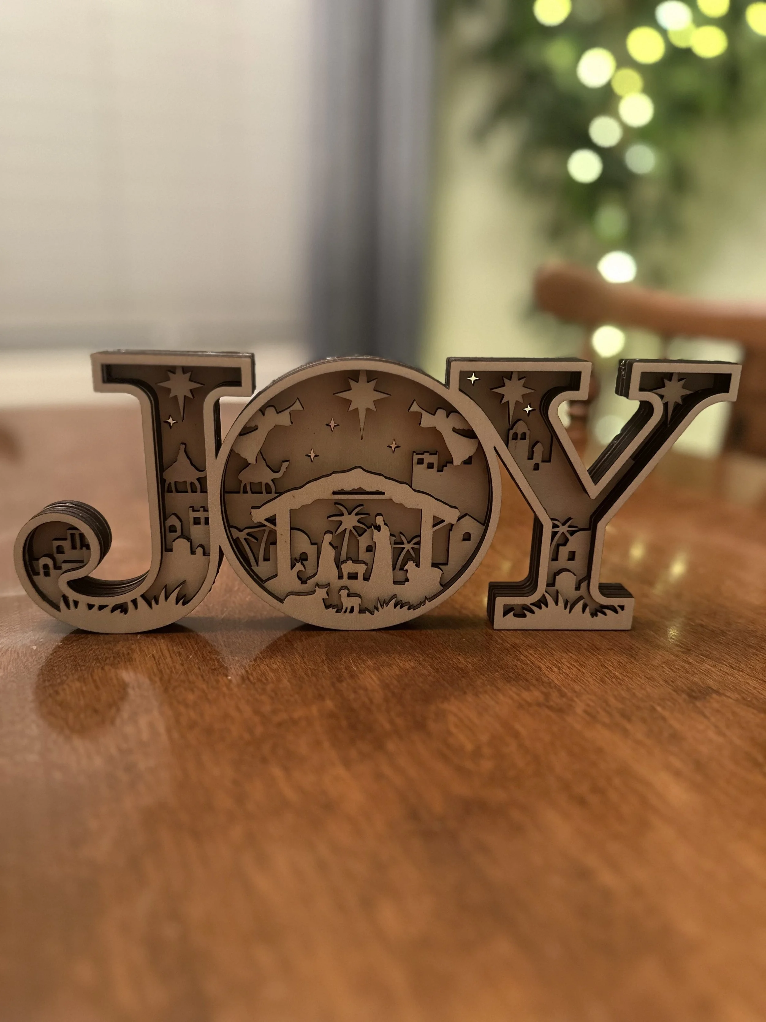 Wood Christmas decoration spelling 'JOY' with nativity scene inside the letters, placed on a wooden table, blurred green background with lights.