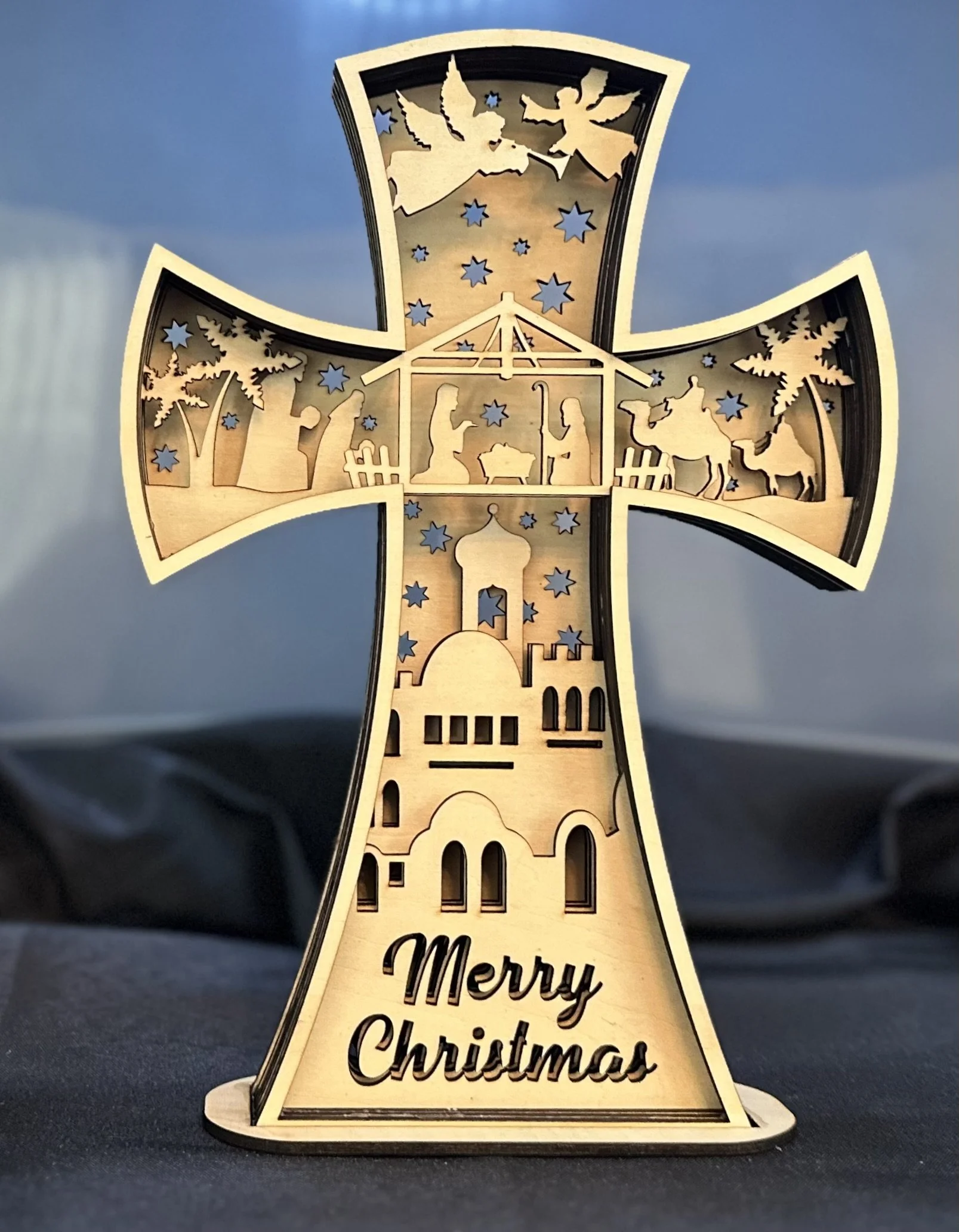 Decorative wooden Christmas cross featuring a nativity scene, angels, and stars, with 'Merry Christmas' inscribed at the base.
