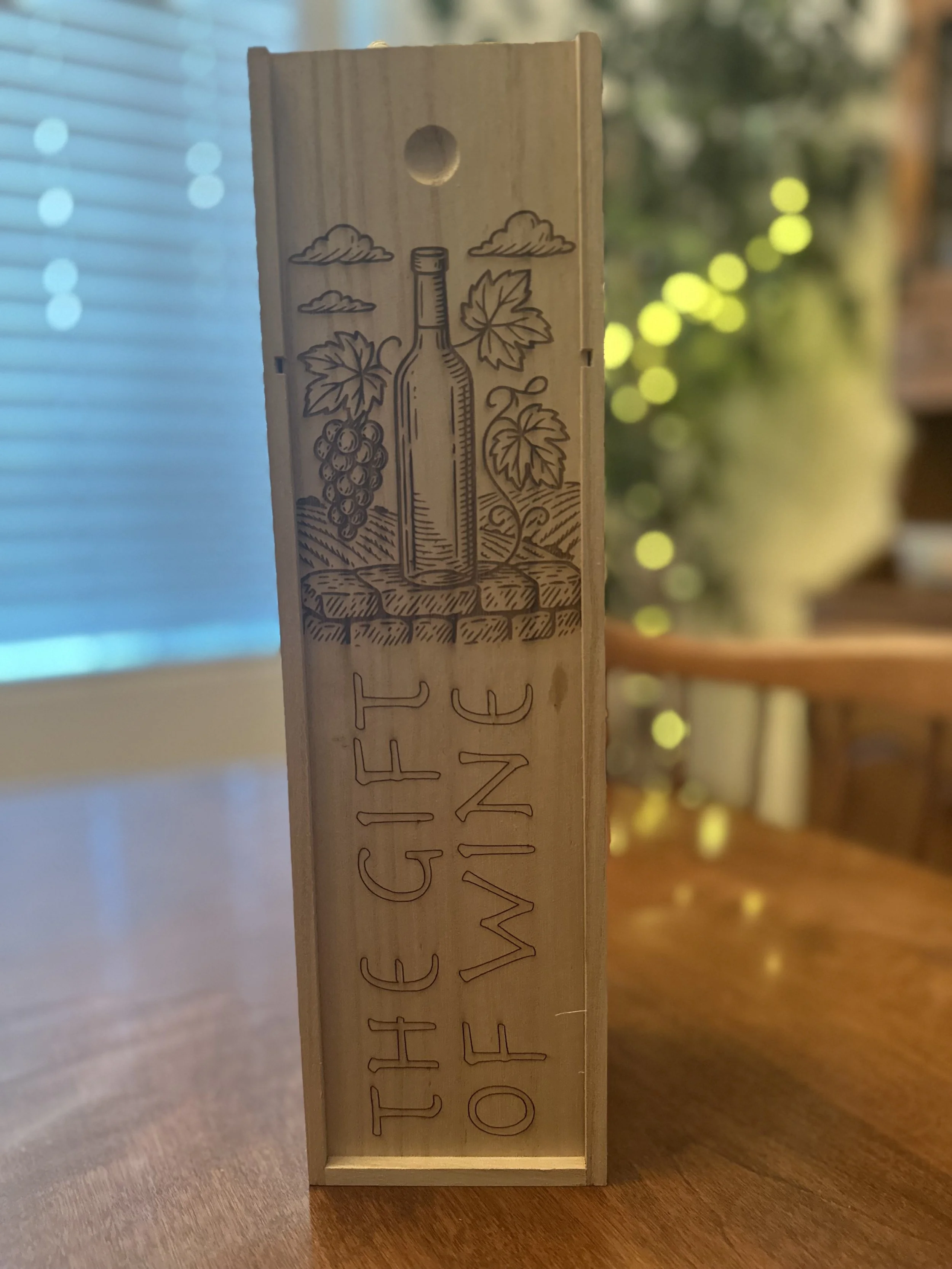 Wooden sign with engraved wine bottle, grapevine leaves, grapes, and the words 'THE GIFT OF WINE'.