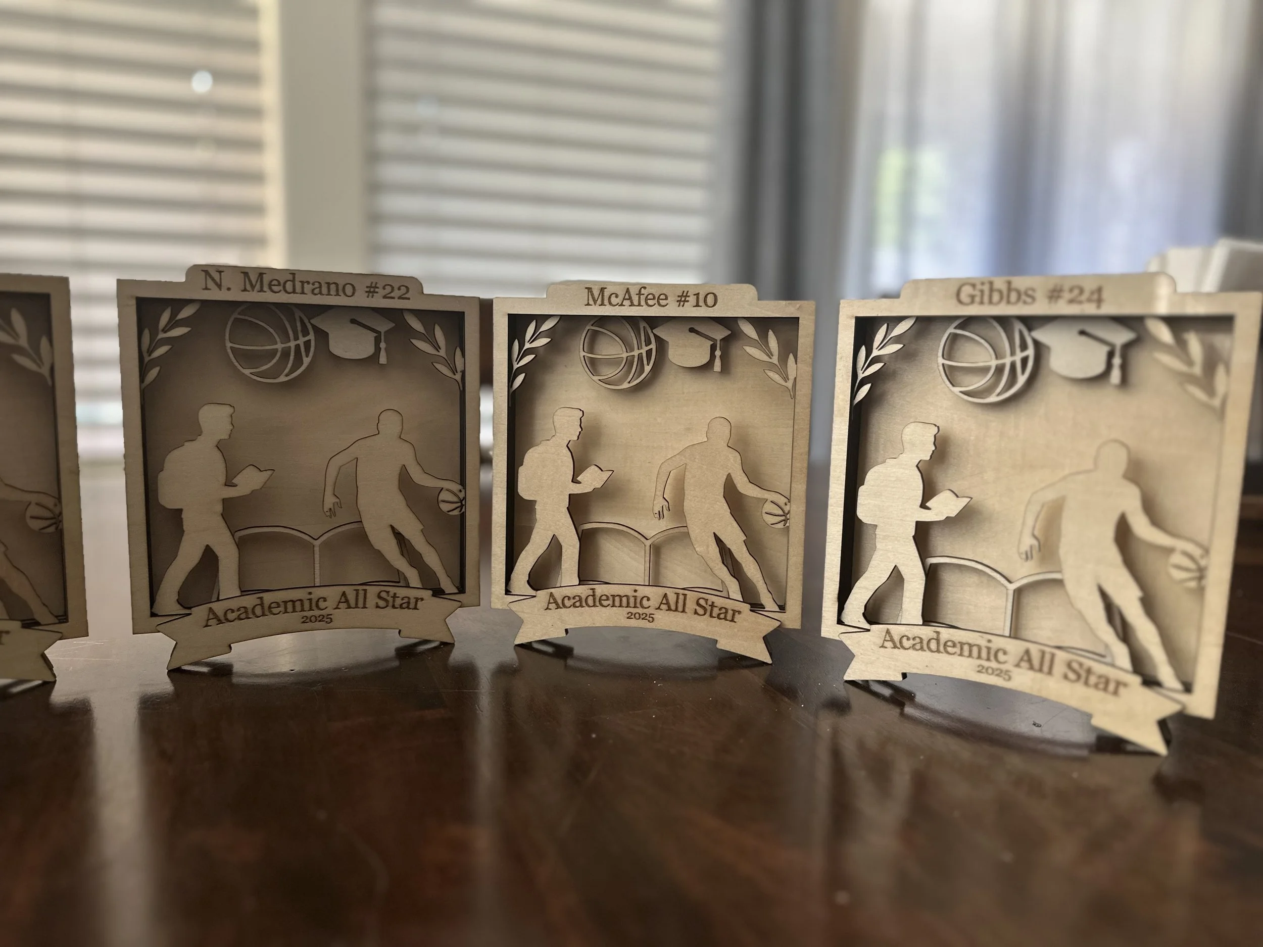 Wooden basketball award plaques with cutout silhouettes of a player dribbling, each engraved with names and numbers, titled "Academic All Star 2025."