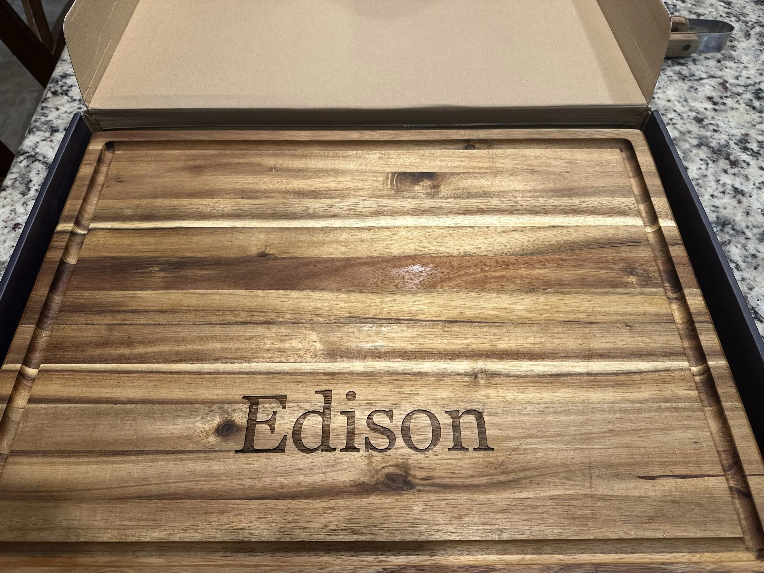 A wooden cutting board with the word "Edison" engraved on it, inside an open cardboard box on a granite countertop.