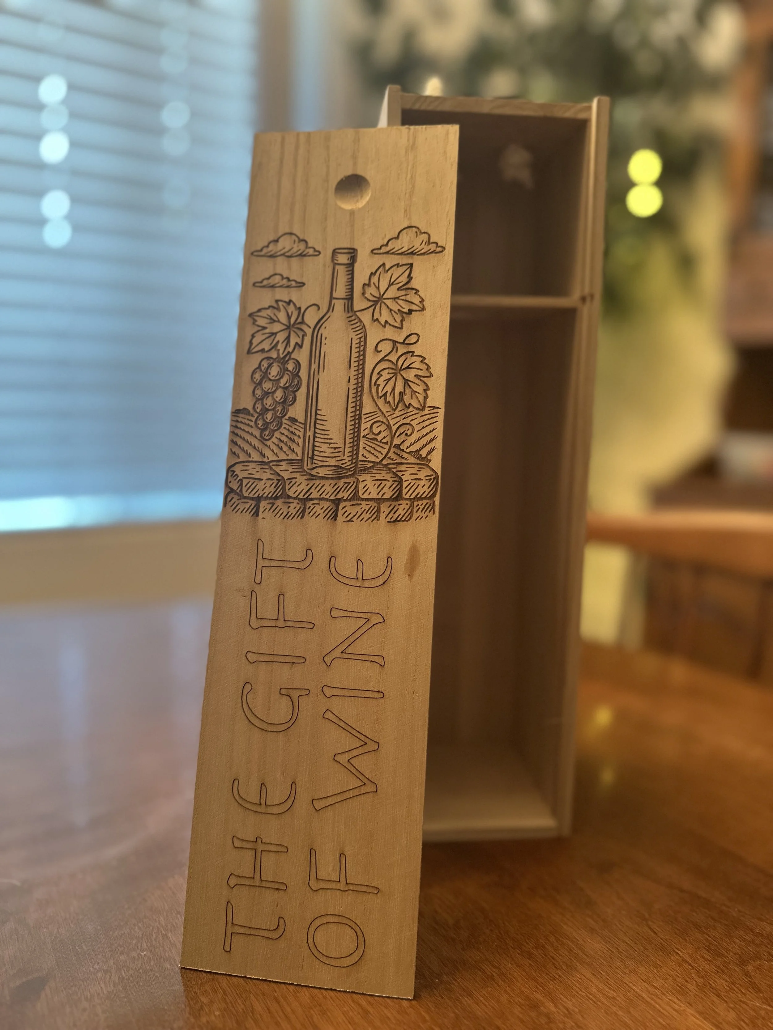 Wooden wine-themed box with engraved illustration of a wine bottle, grapevine, and clouds, and text that reads 'THE GIFT OF WINE.'
