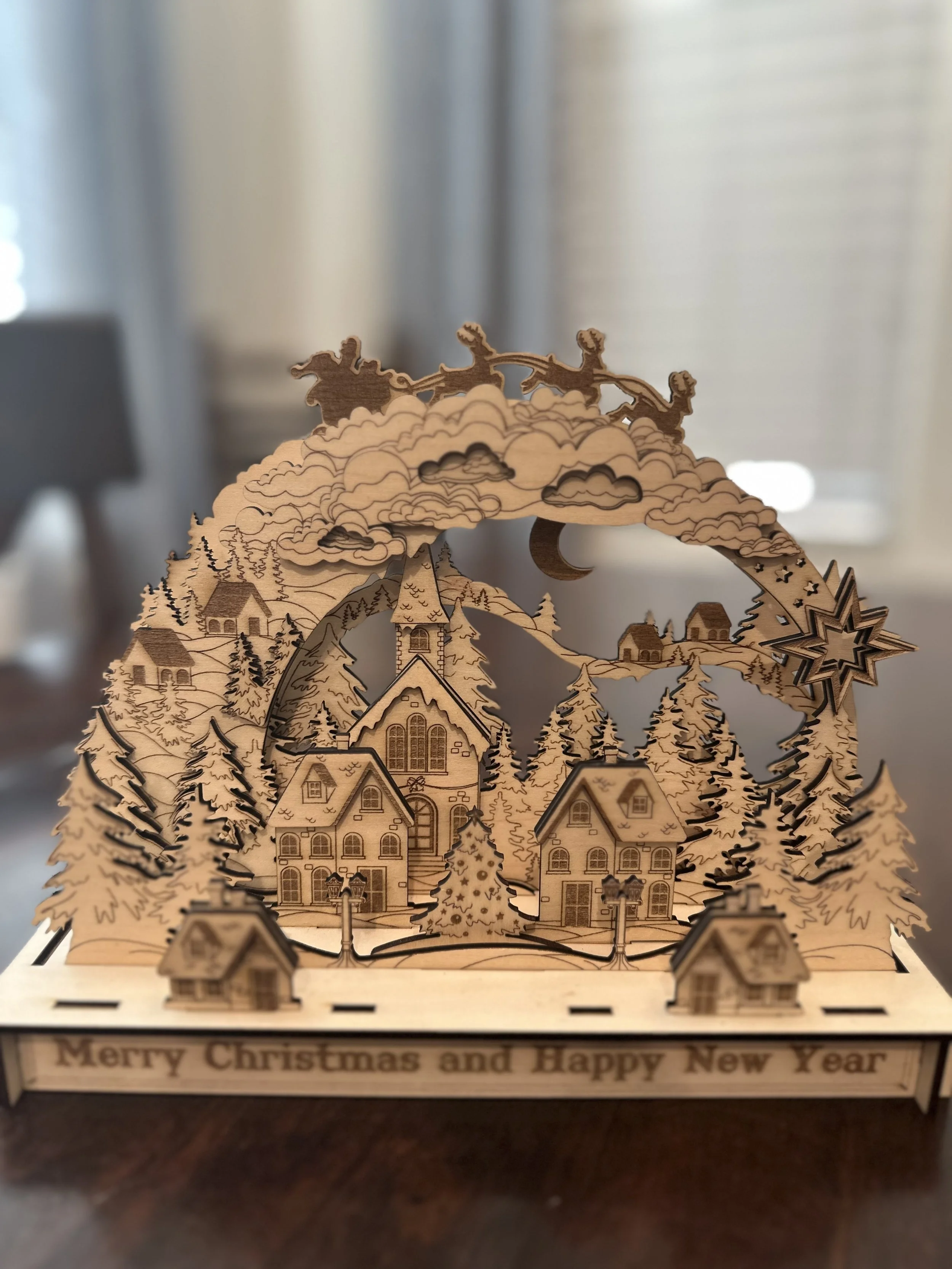 Wooden Christmas and New Year holiday decoration featuring houses, trees, clouds, and a star with the message 'Merry Christmas and Happy New Year' at the base.