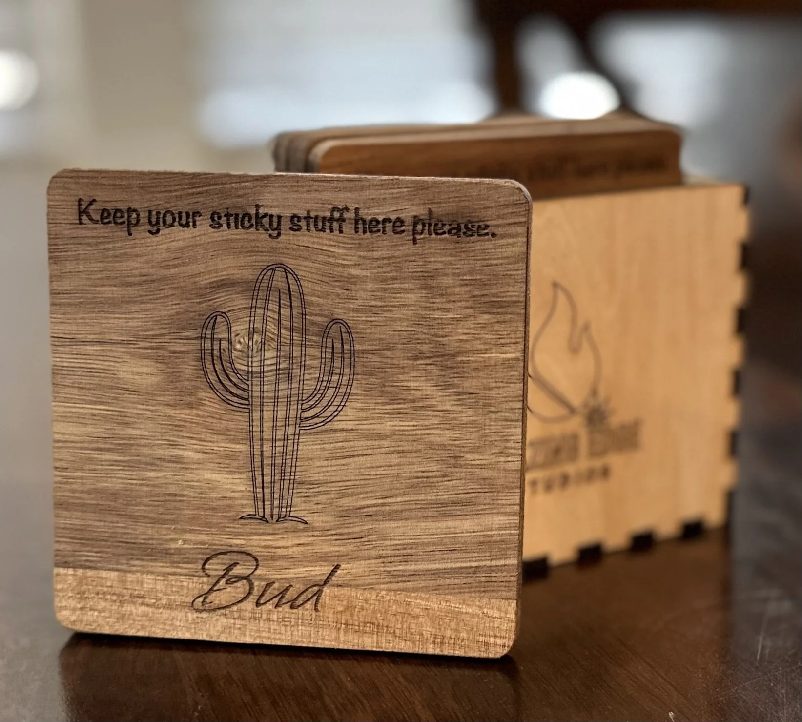A wooden coaster with a drawing of a cactus, the text 'Keep your sticky stuff here please,' and the name 'Bud' written at the bottom.