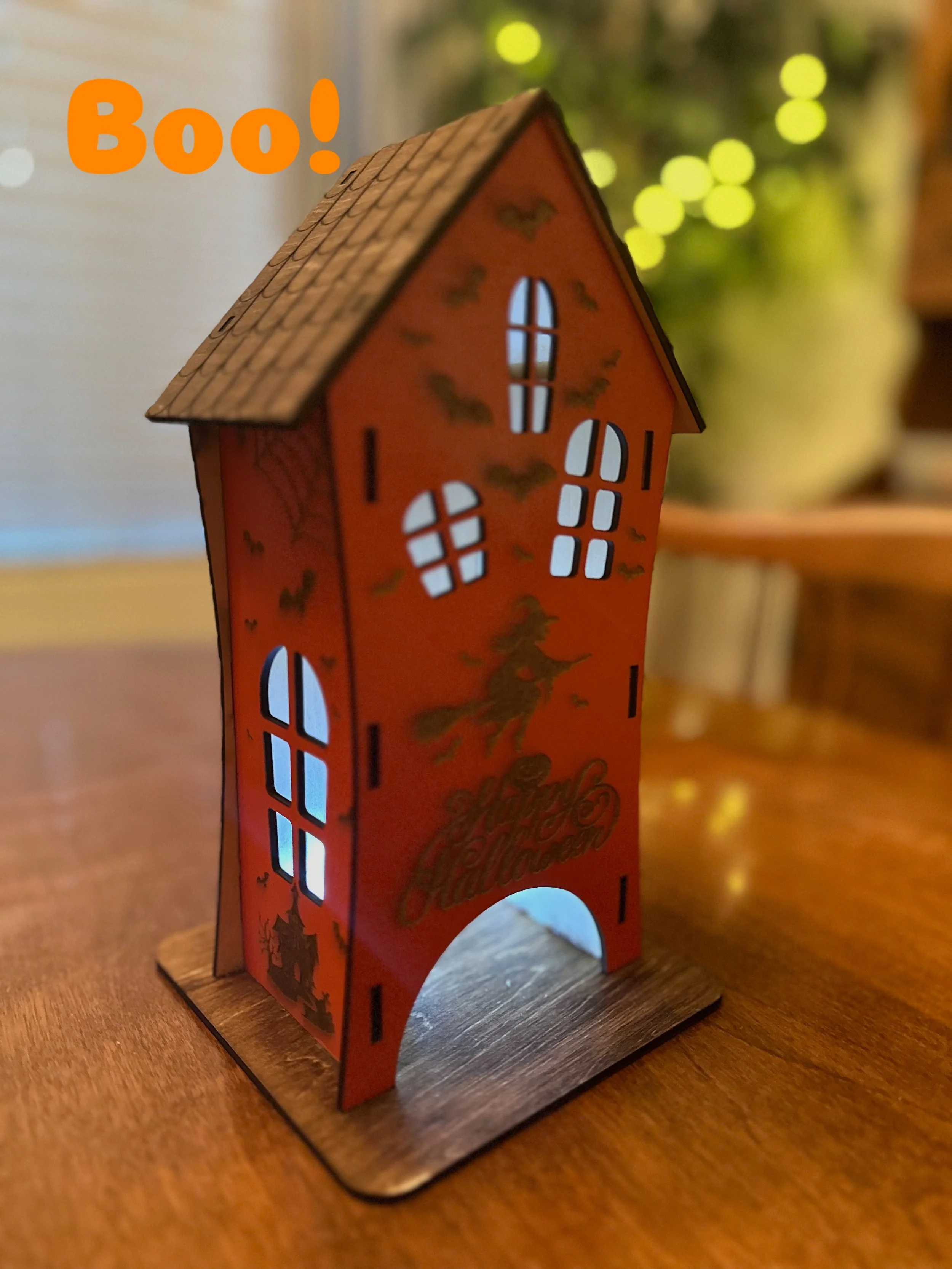 Small orange Halloween decoration house with windows and a front arch, featuring witch and bat illustrations and the words 'Happy Halloween', placed on a wooden surface.