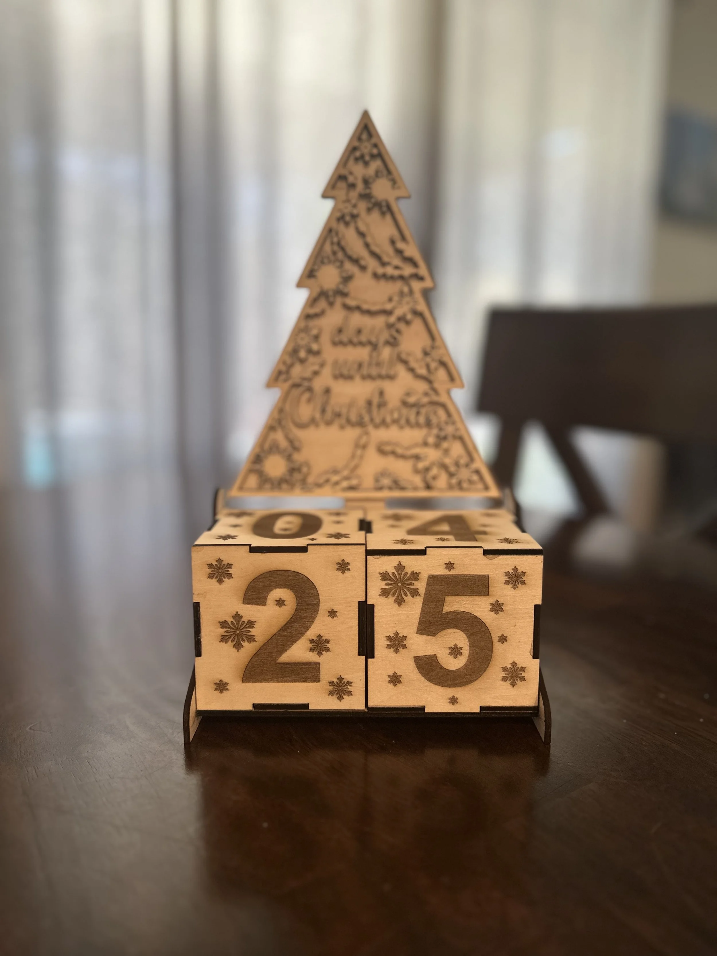 Wooden advent calendar with numbered cubes displaying '25' in the foreground and a Christmas tree-shaped decoration in the background.