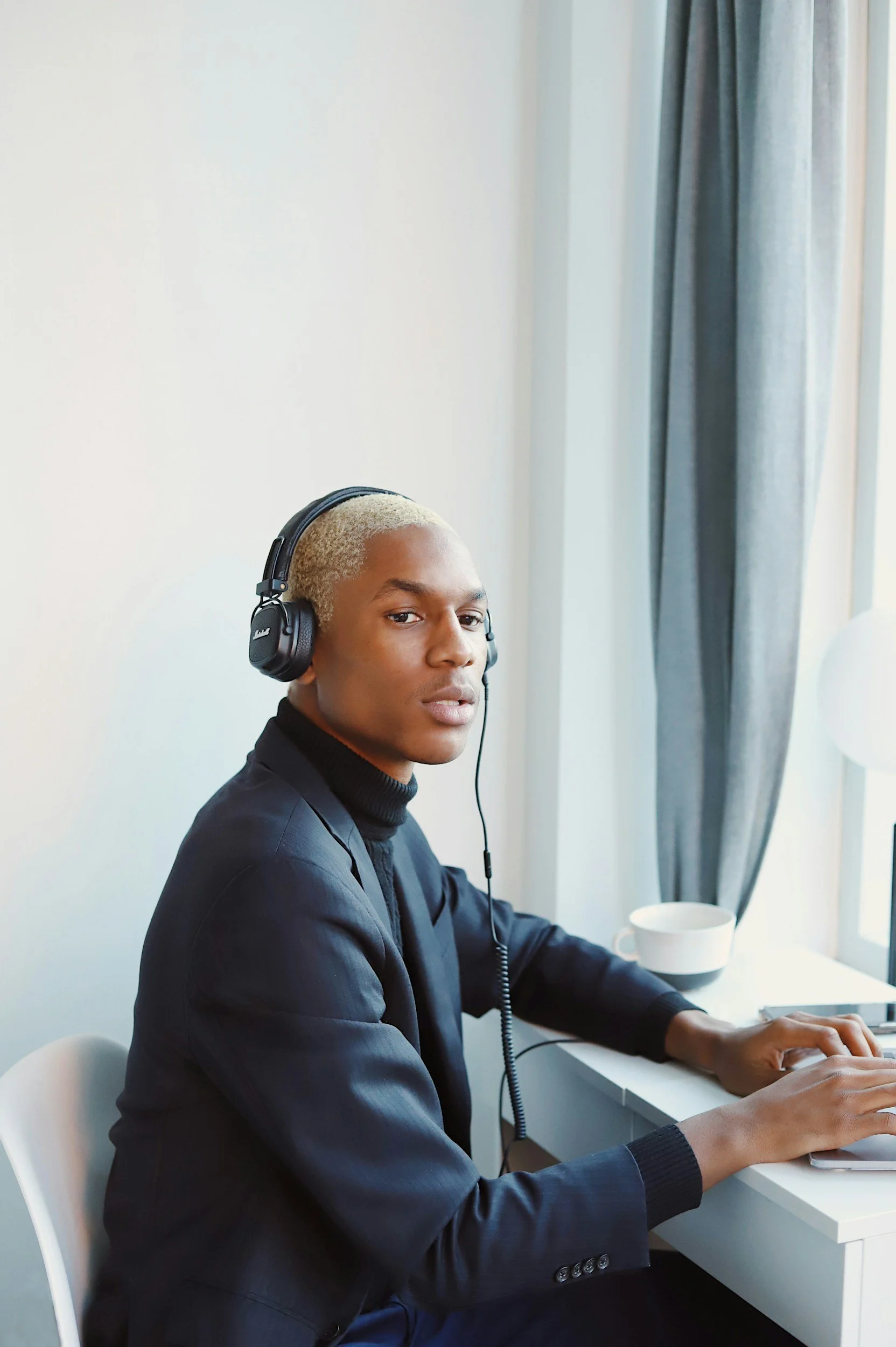 Young person with bleached blonde hair wearing headphones, working on a laptop at a white desk in a bright room with gray curtains.