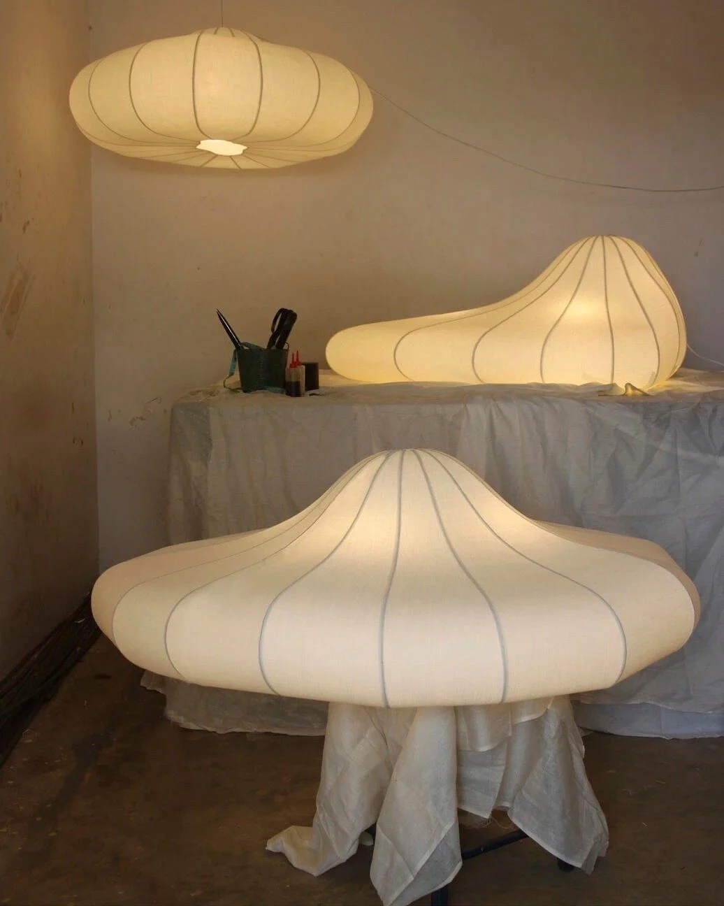 Three cloud-shaped lamps emitting warm light, placed on a table covered with a white cloth, in a room with plain walls and some tools and supplies visible on the table.