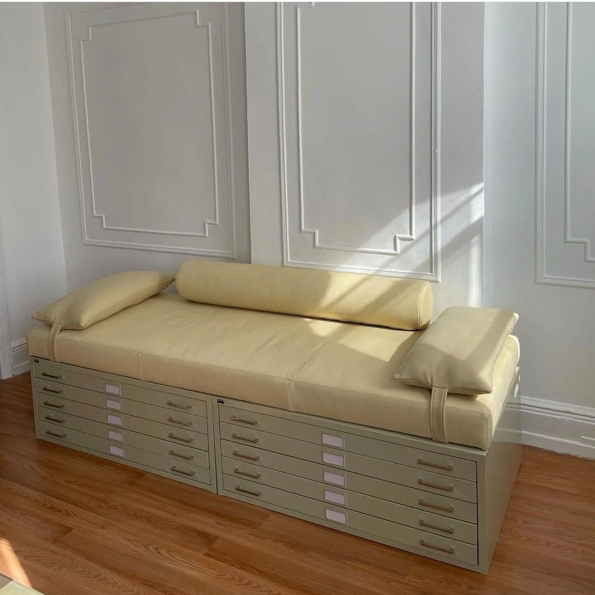 A vintage daybed with a light yellow mattress and two matching cushions, placed against a white wall with decorative molding. The base of the daybed features multiple small drawers, and the room has wooden flooring and sunlight casting shadows on the