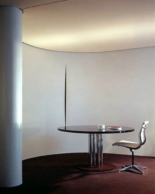 Empty room with a round table, a chair, a white wall, and a ceiling with hidden lighting.