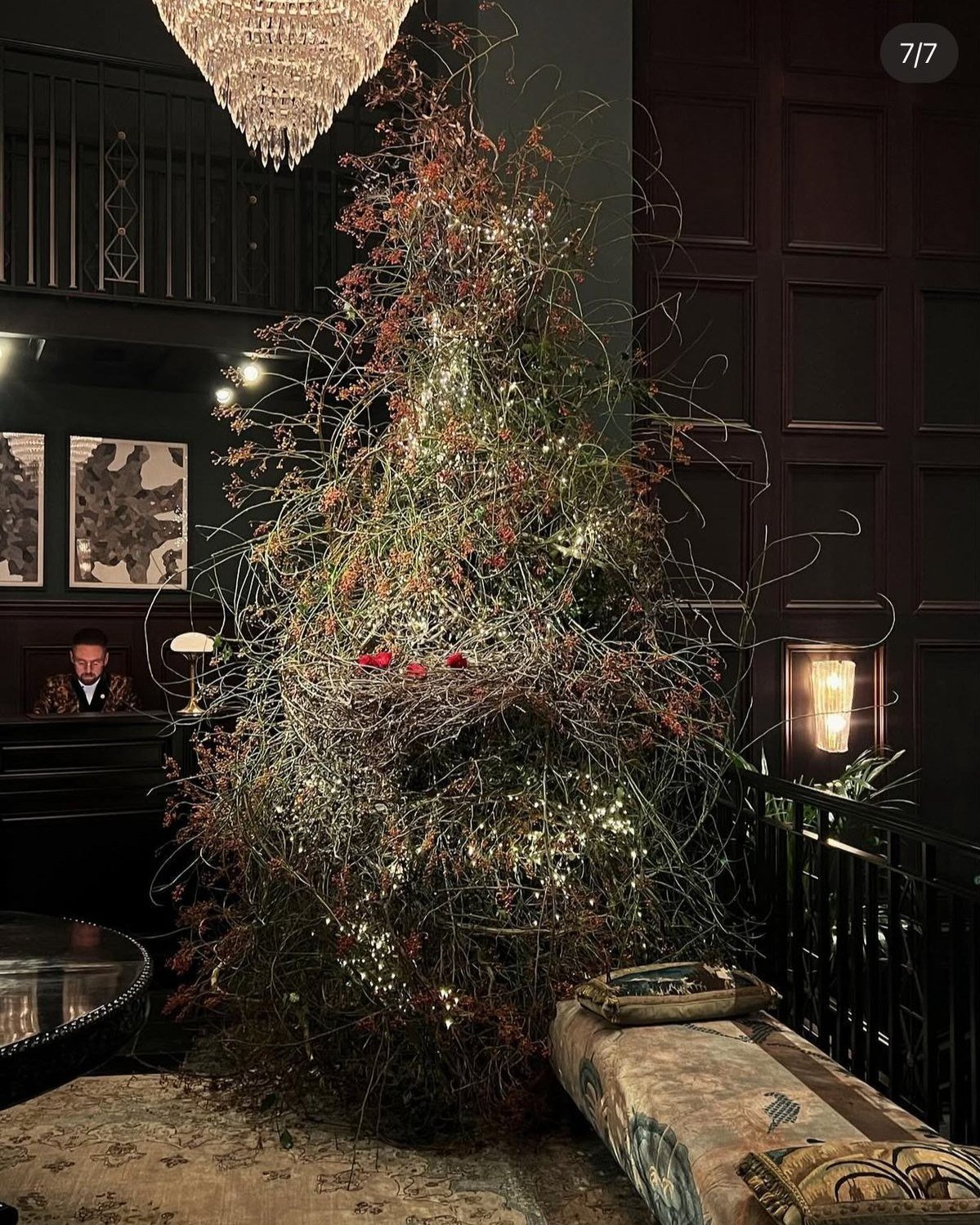 A large, artistic Christmas tree decorated with lights and minimal adornments in a dark, elegant room with wood-paneled walls and framed artwork. A velvet-upholstered seating area is nearby, with a man standing behind a reception desk in the backgrou