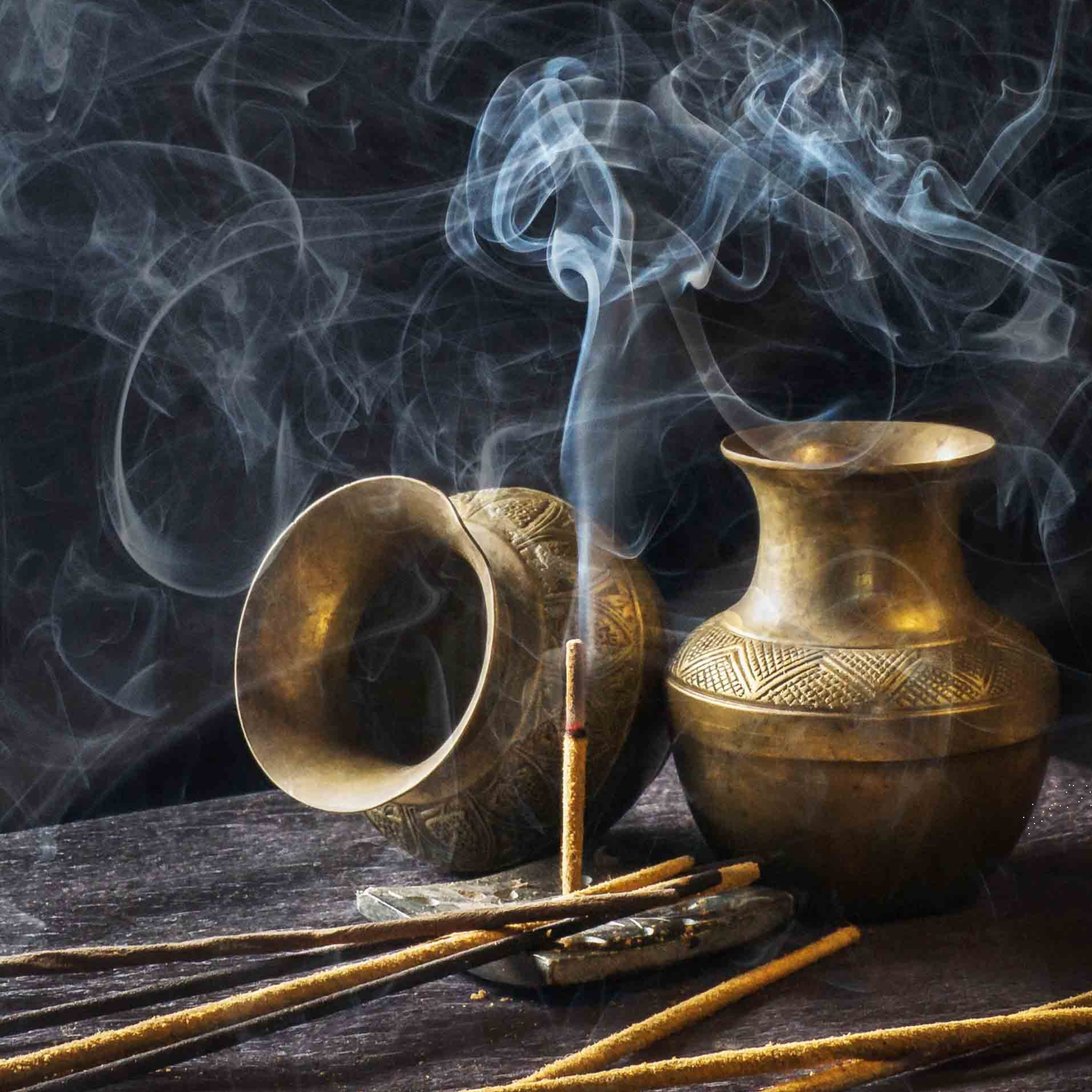 Two brass or bronze incense holders emitting smoke, placed on a dark wooden surface, with several lit incense sticks and additional incense holders in the background.