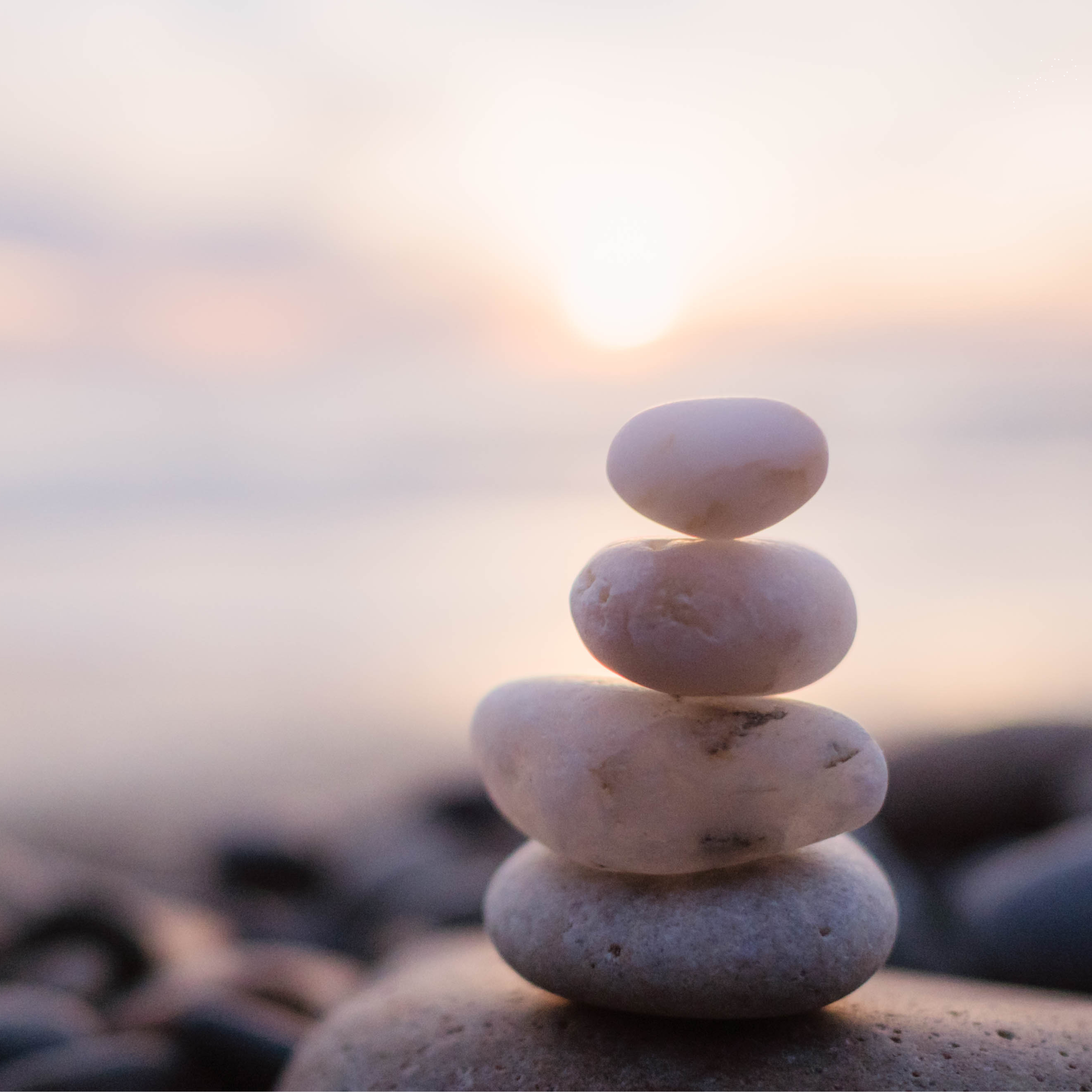 Stack of five smooth, white and beige stones balanced on top of each other on a beach with a blurred background of water and sky at sunset