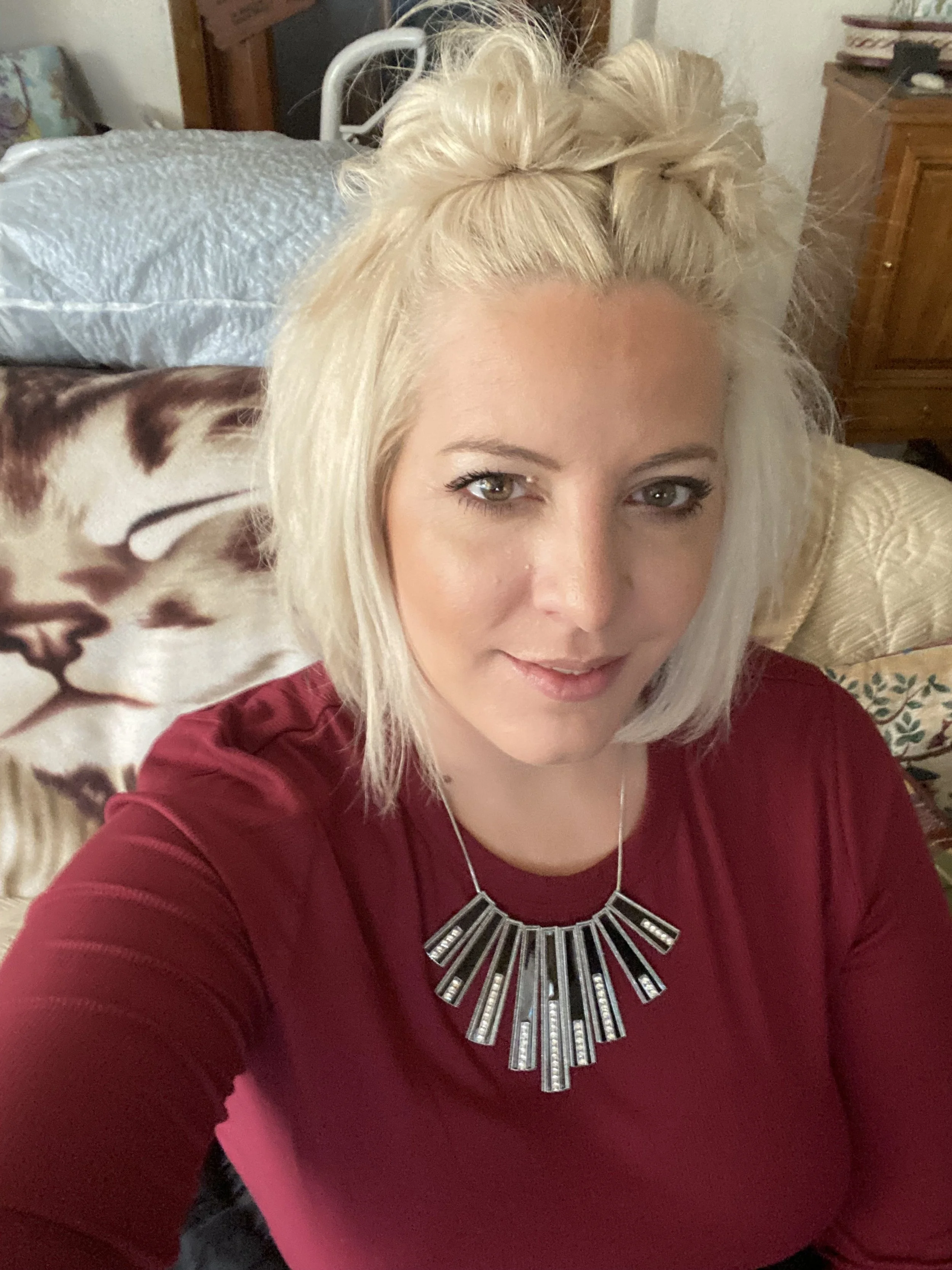 A woman with blonde hair styled in a half-up, half-down fashion, wearing a burgundy top and a statement necklace with geometric shapes, taking a selfie in a room with a bed, pillows, and wooden furniture in the background.