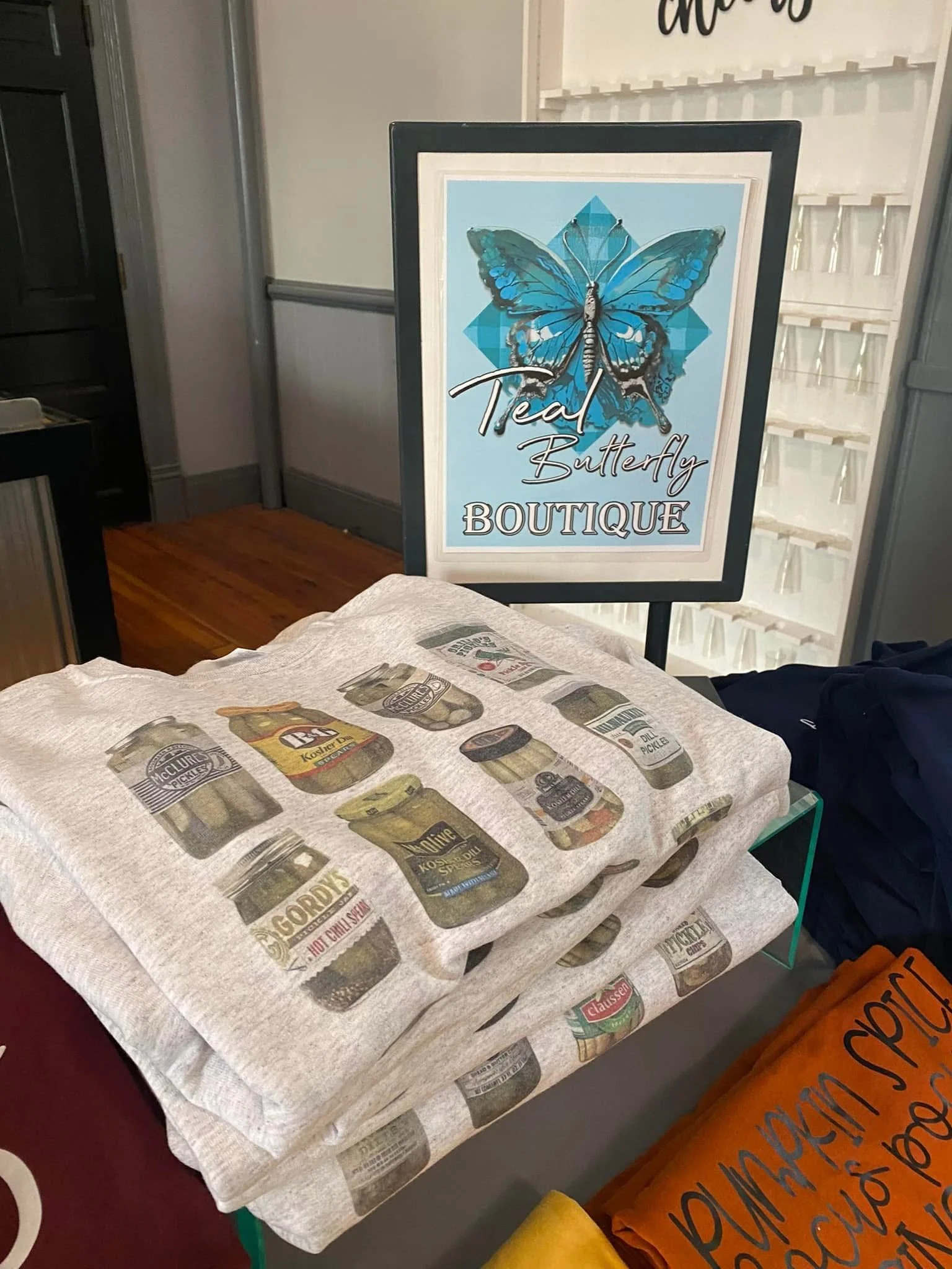 Display stand with folded fabric featuring images of various pickles, next to a sign for Teal Butterfly Boutique, which has a blue butterfly logo.
