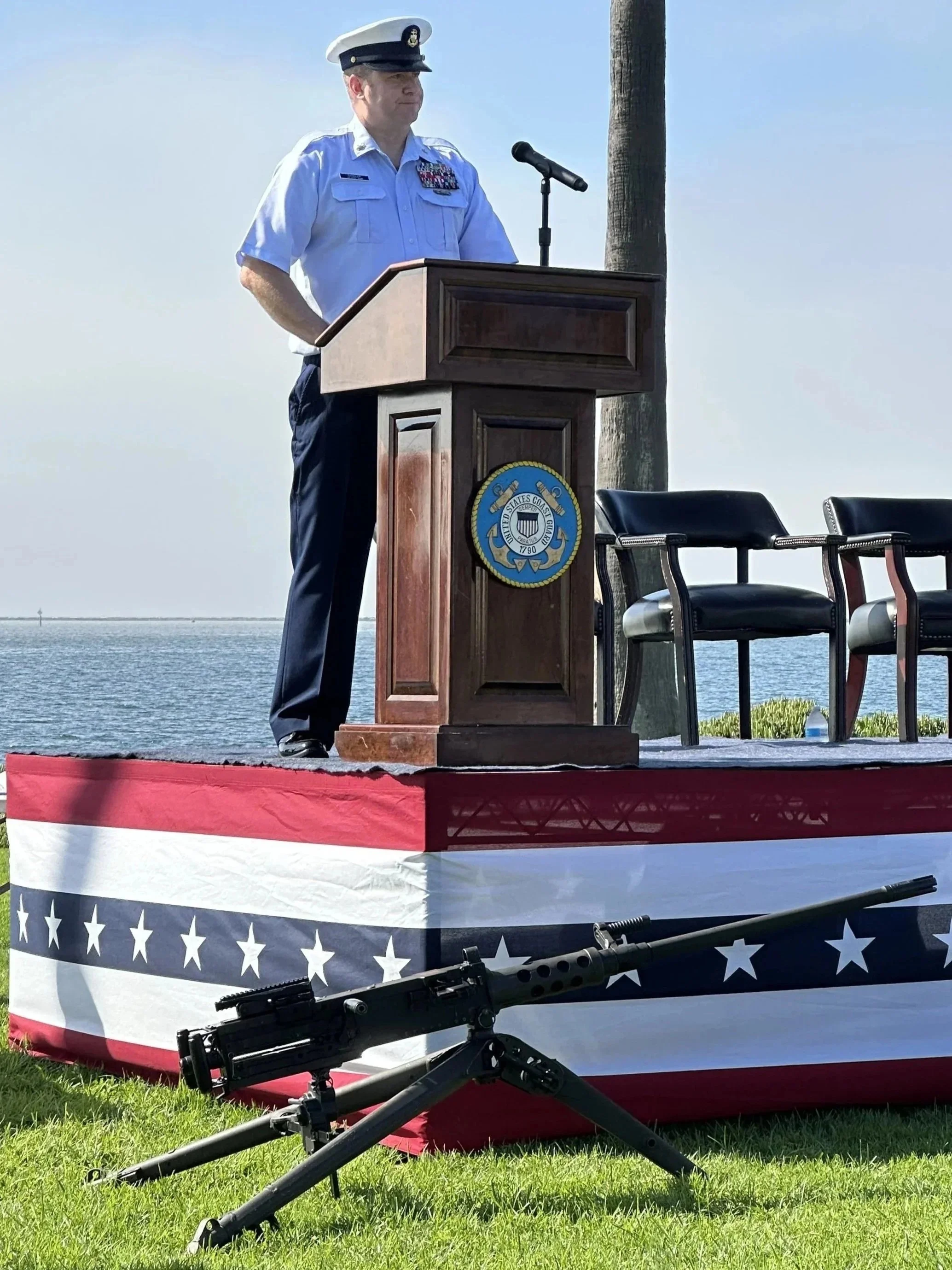 A man in a United States Coast Guard uniform stands at a podium during a ceremony near the water, with a machine gun and a red, white, and blue patriotic display in the foreground.