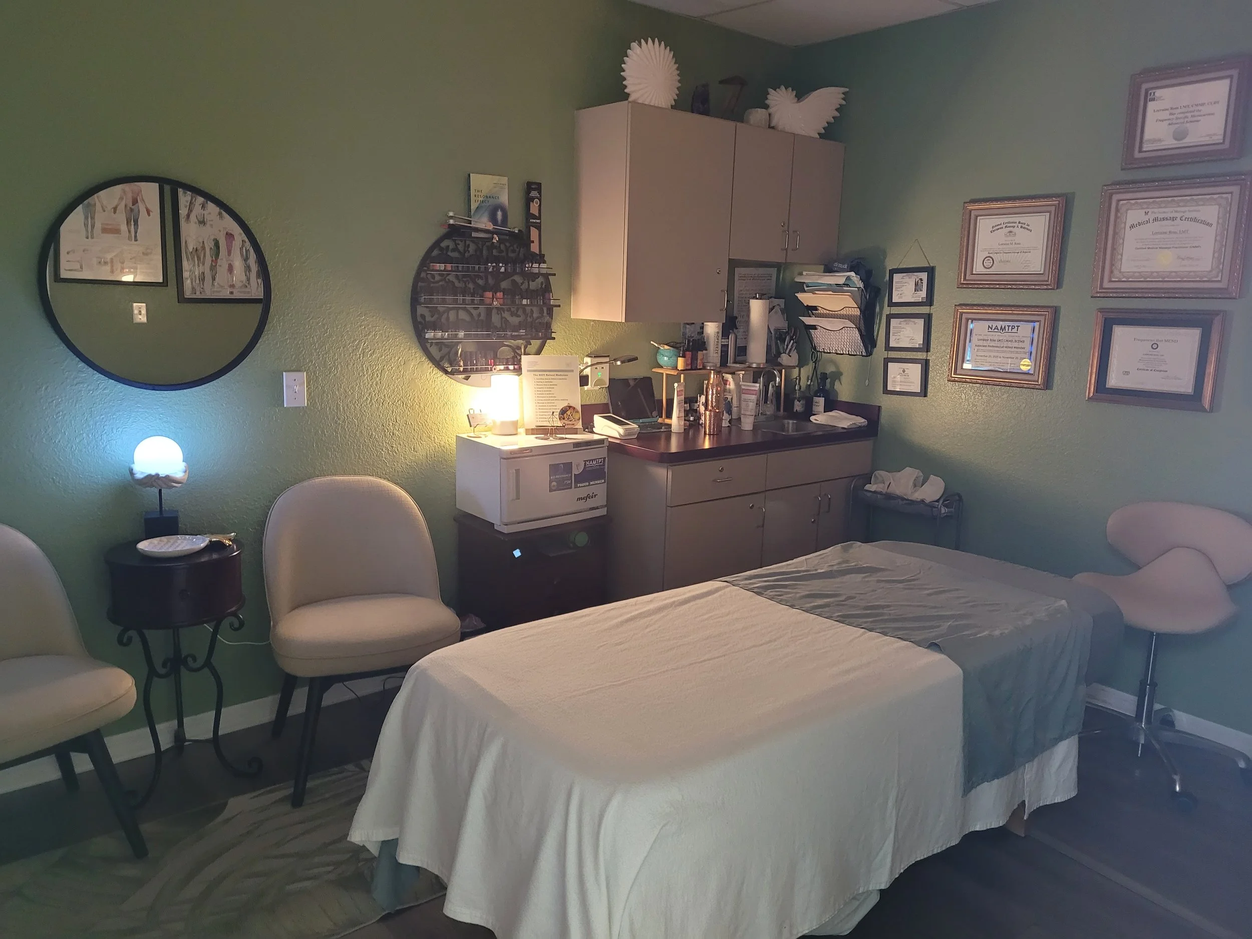A massage therapy room with a massage table covered in a beige sheet, framed certificates and wall art.