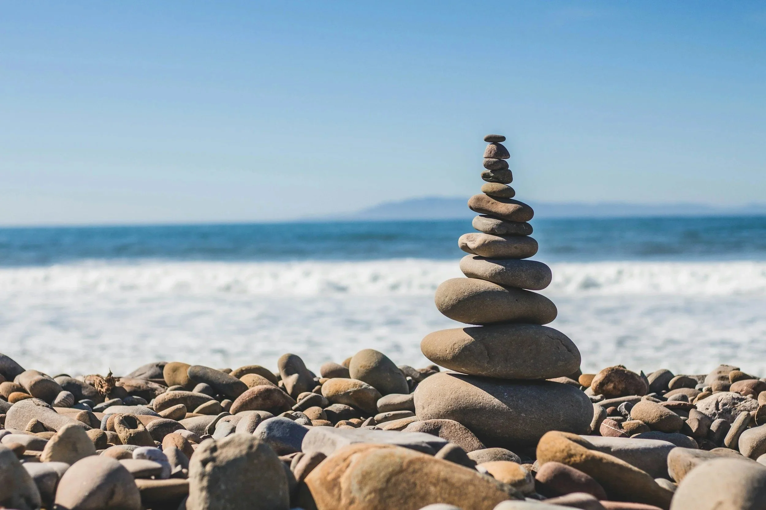 A stack of smooth, flat stones balanced on top of each other on a rocky beach with ocean waves and a distant landmass in the background.