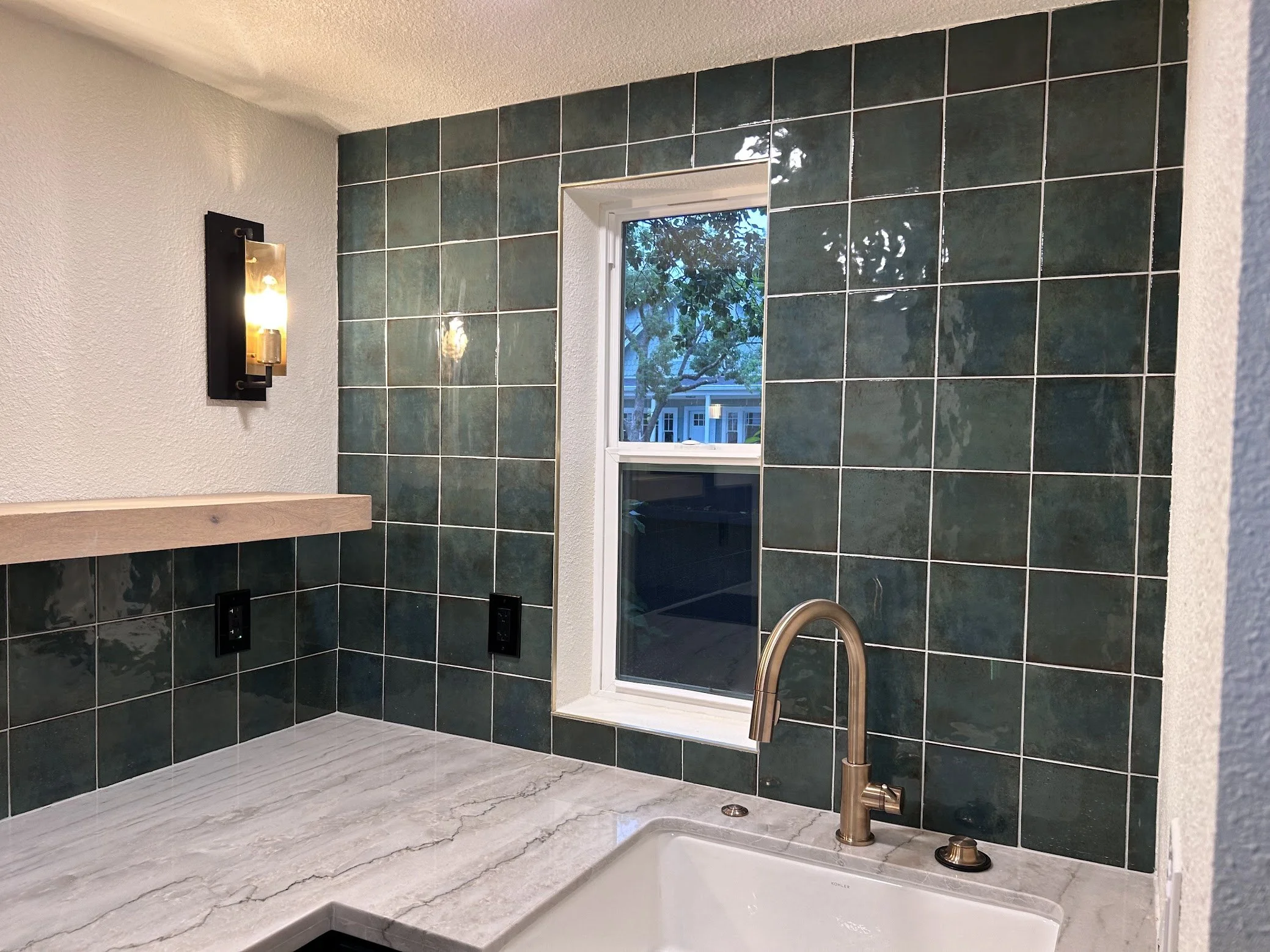 A kitchen or bathroom corner with dark green tiled wall, a small window overlooking trees and houses, a marble countertop with a sink and a brass faucet, a wooden shelf, a wall sconce, and two electrical outlets.