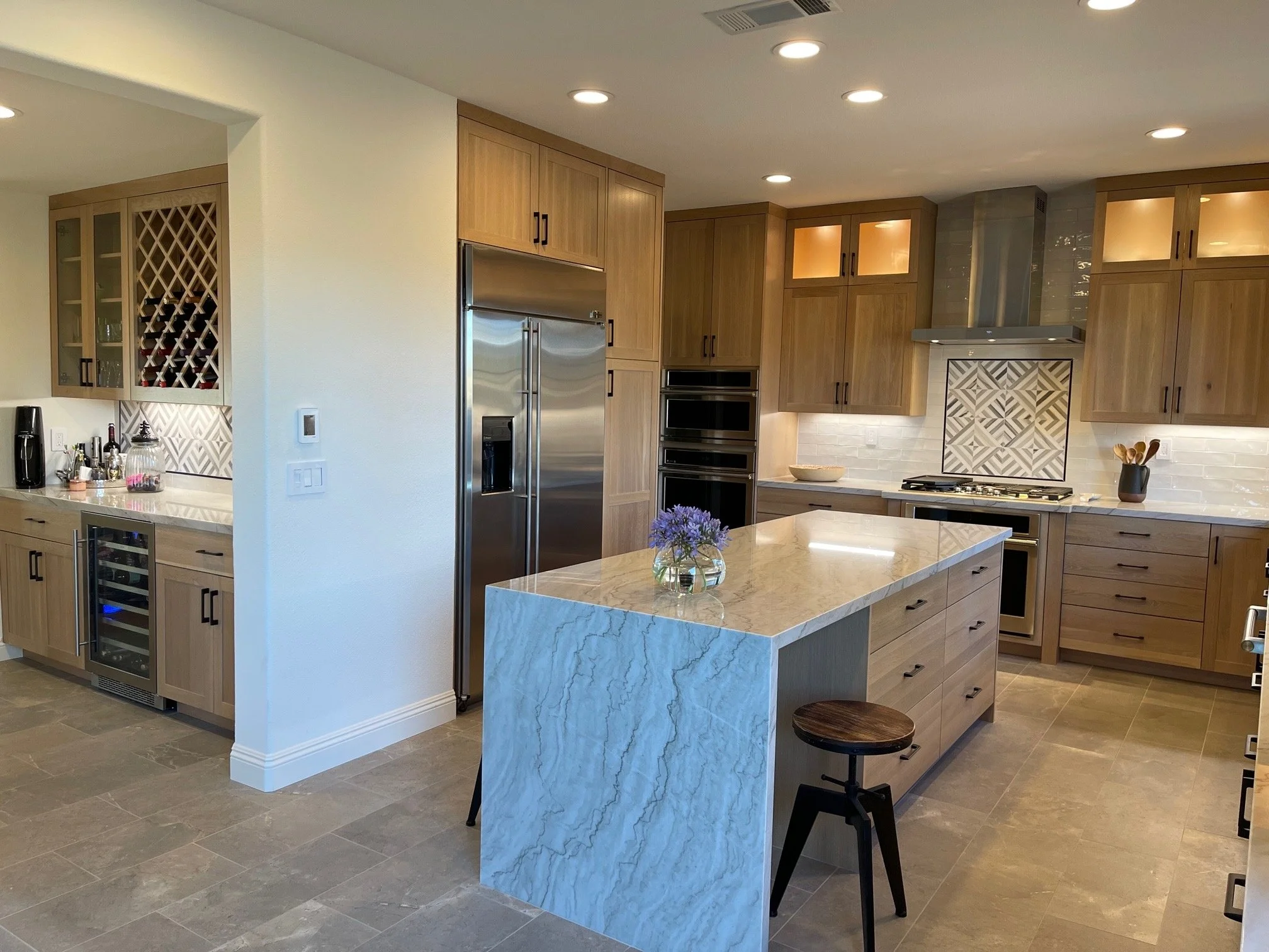 Modern kitchen with wooden cabinets, stainless steel appliances including double ovens and a refrigerator, a marble kitchen island with a small flower arrangement, and a stool.
