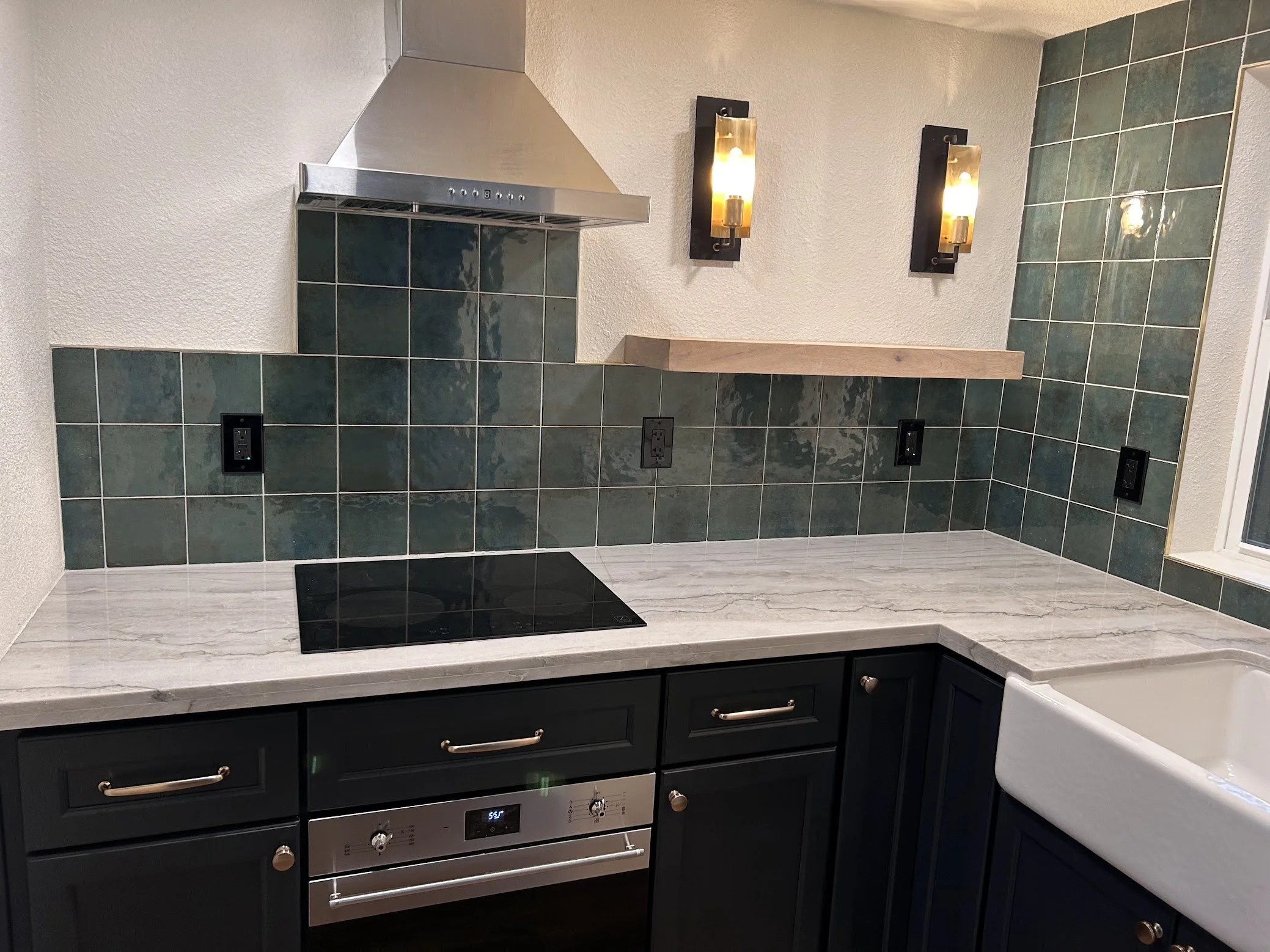 Kitchen with black lower cabinets, marble countertop, green tiled backsplash, stainless steel range hood, black electric cooktop, wall-mounted light fixtures, and a white farmhouse sink.