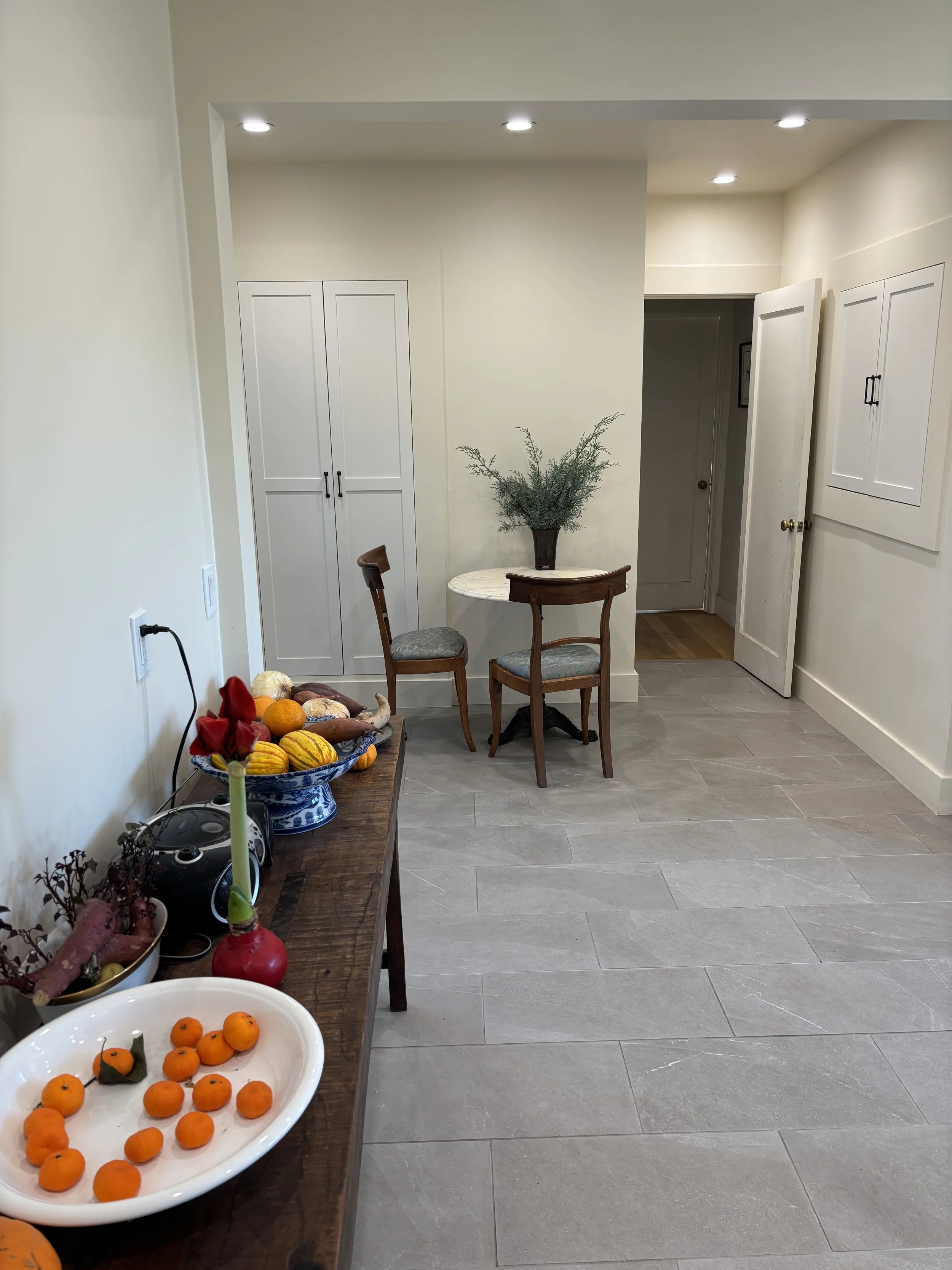Interior view of a home dining area with a wooden table, two chairs, a potted plant, and a cabinet. A sideboard on the left holds decorative gourds, a bowl of small oranges, and a candlestick with a yellow candle. The room has recessed lighting and l