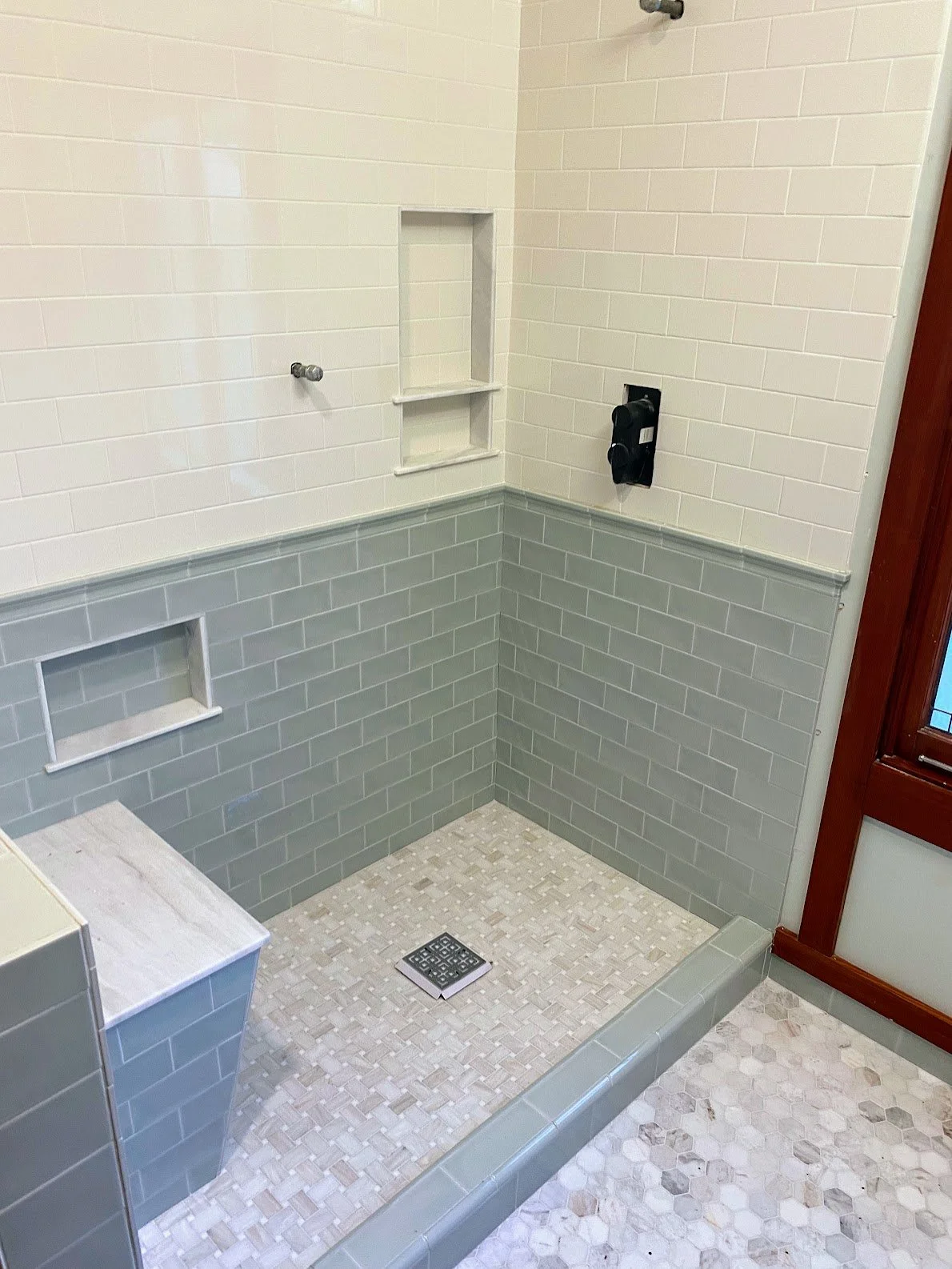 Empty walk-in shower area with tiled walls and floor, built-in shelves, drain on the floor, and a black shower faucet with a wooden-framed window nearby.