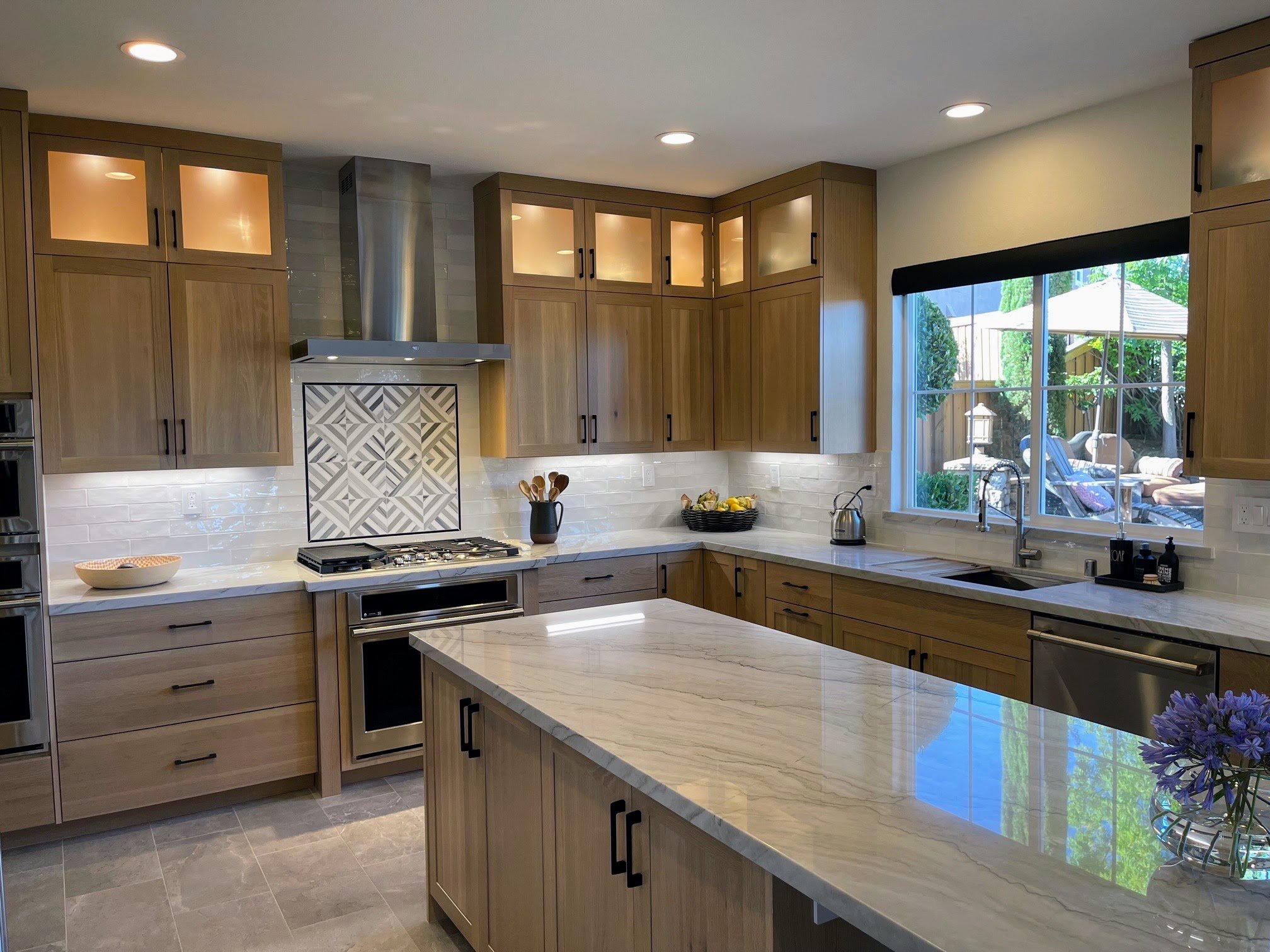 Modern kitchen with wooden cabinets, white marble countertops, and a large window overlooking an outdoor patio with chairs and greenery.