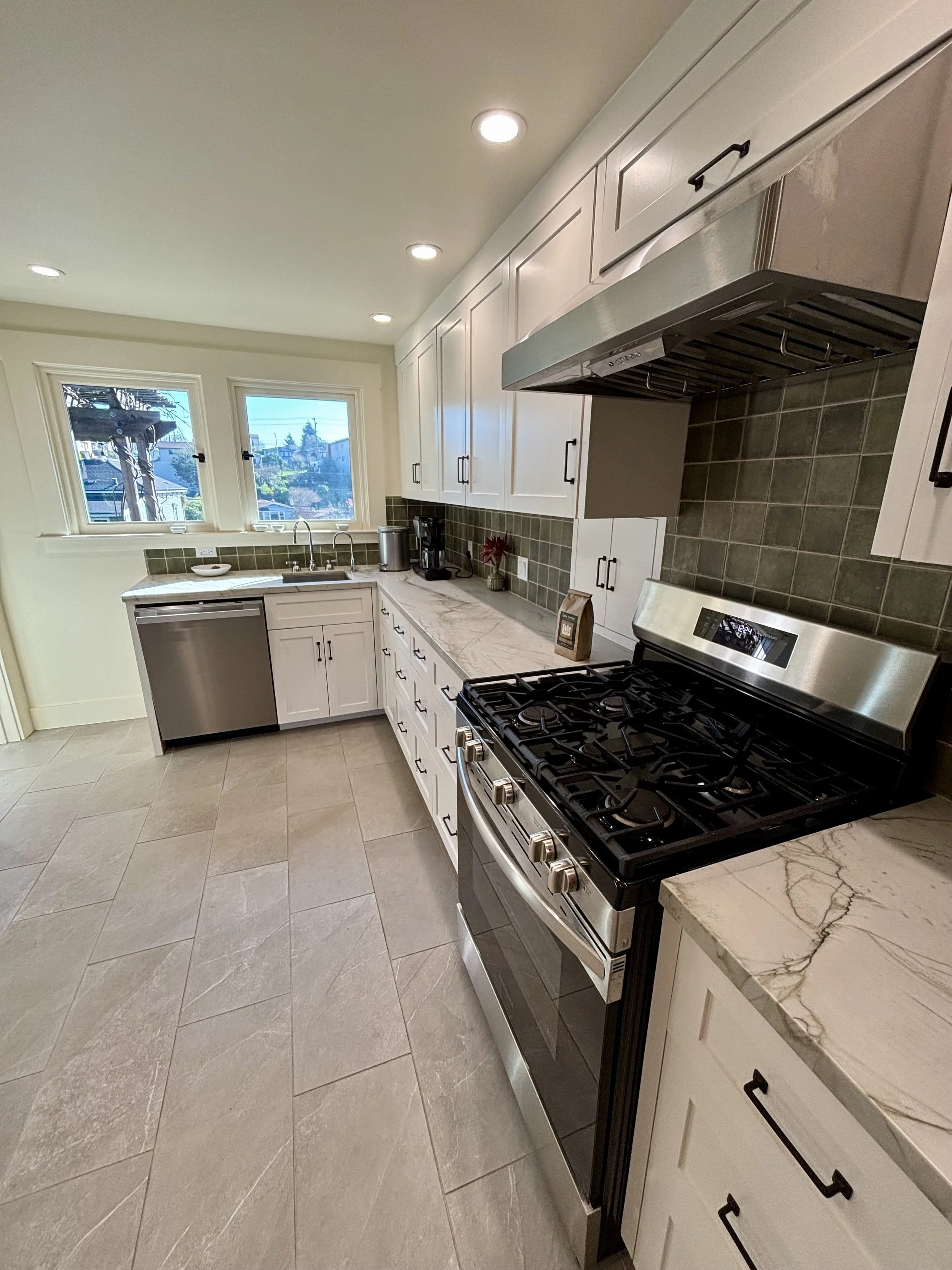 Modern kitchen with white cabinets, stainless steel appliances, green tiled backsplash, and beige tiled floor, illuminated by ceiling lights and natural light from two windows.