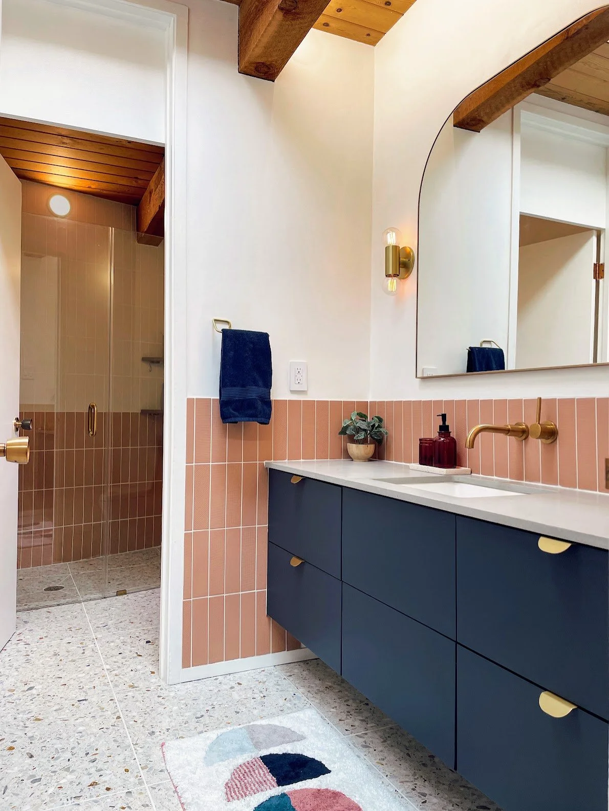 Modern bathroom with pink tiles, navy blue vanity with yellow handles, a large mirror, a brass wall-mounted light fixture, a plant and soap dispenser on the counter, a glass shower with a gold handle, and a white and multicolored rug on terrazzo floo