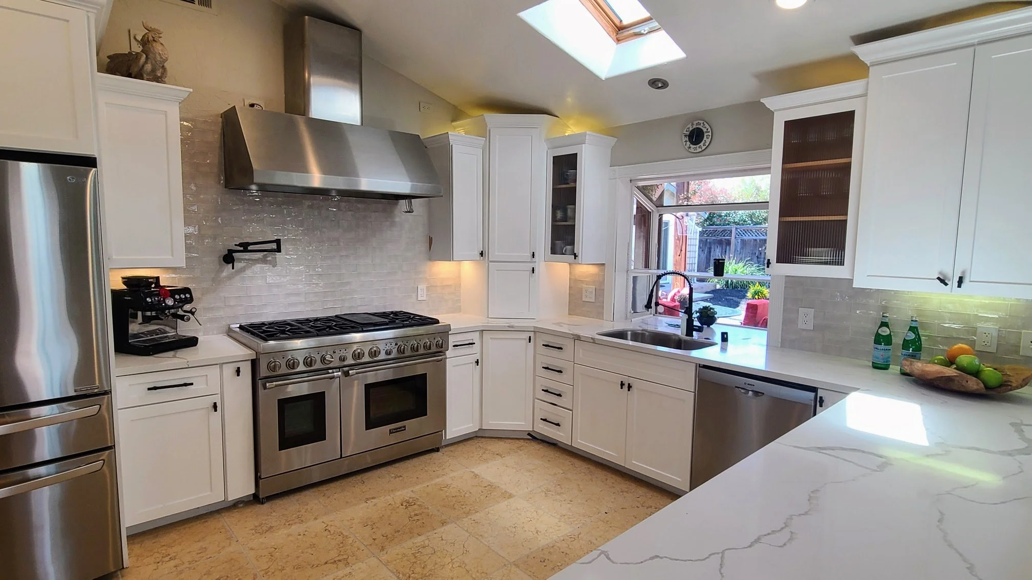 Modern kitchen with white cabinets, stainless steel appliances, a large gas stove and oven, a coffee machine, a dishwasher, a sink beneath a window, white marble countertops, and a skylight.