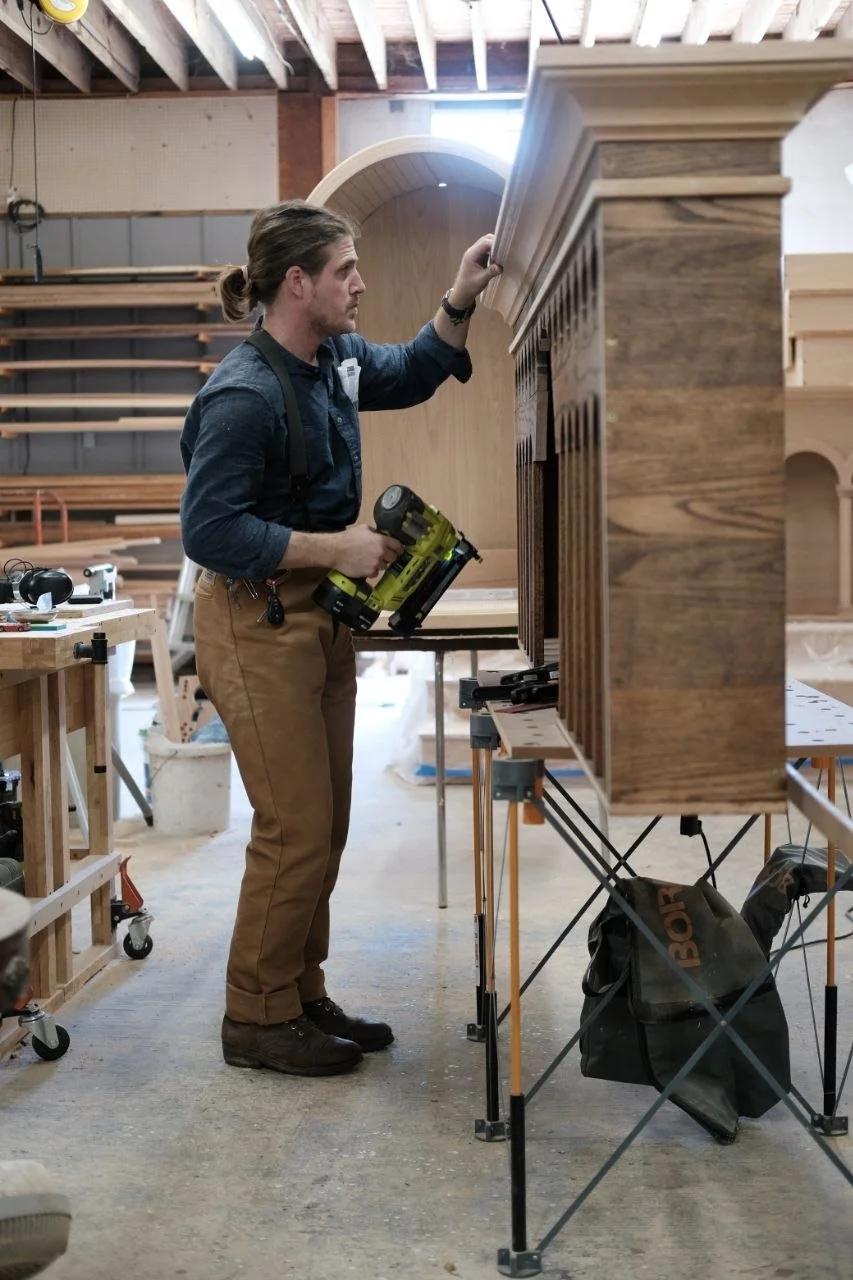 A man with long hair tied back, wearing a dark shirt and tan pants, is working on assembling a wooden structure in a woodworking workshop. He is holding a cordless drill and inspecting the upper part of the wooden piece.