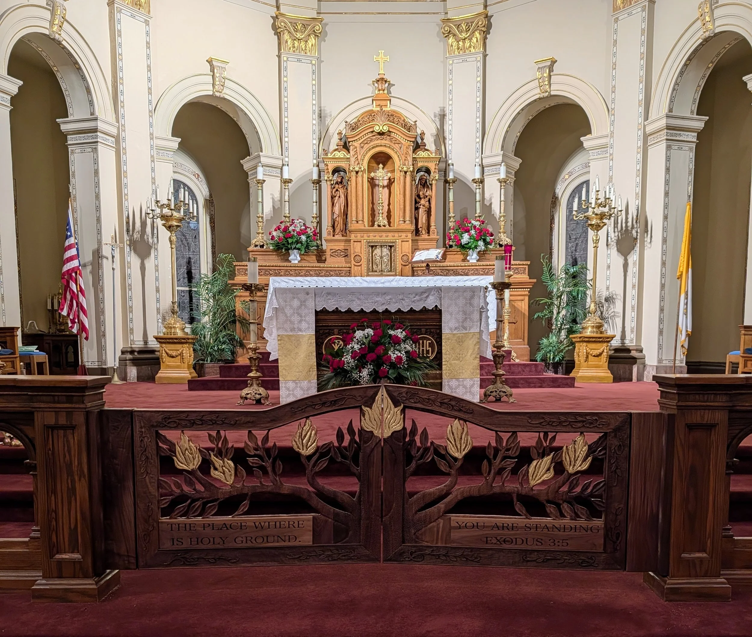 Interior of a church altar with wooden carvings, flowers, candlesticks, and a cross at the center. There are also American flags and a yellow flag on either side of the altar. The altar is decorated with a lace cloth, and there is a Bible resting on a stand. The front of the altar has a wooden panel with the inscription: "The place where the Lord is holy ground," and a biblical quote from Exodus 3:5, "You are standing on holy ground." The background features ornate architectural details and arched niches.