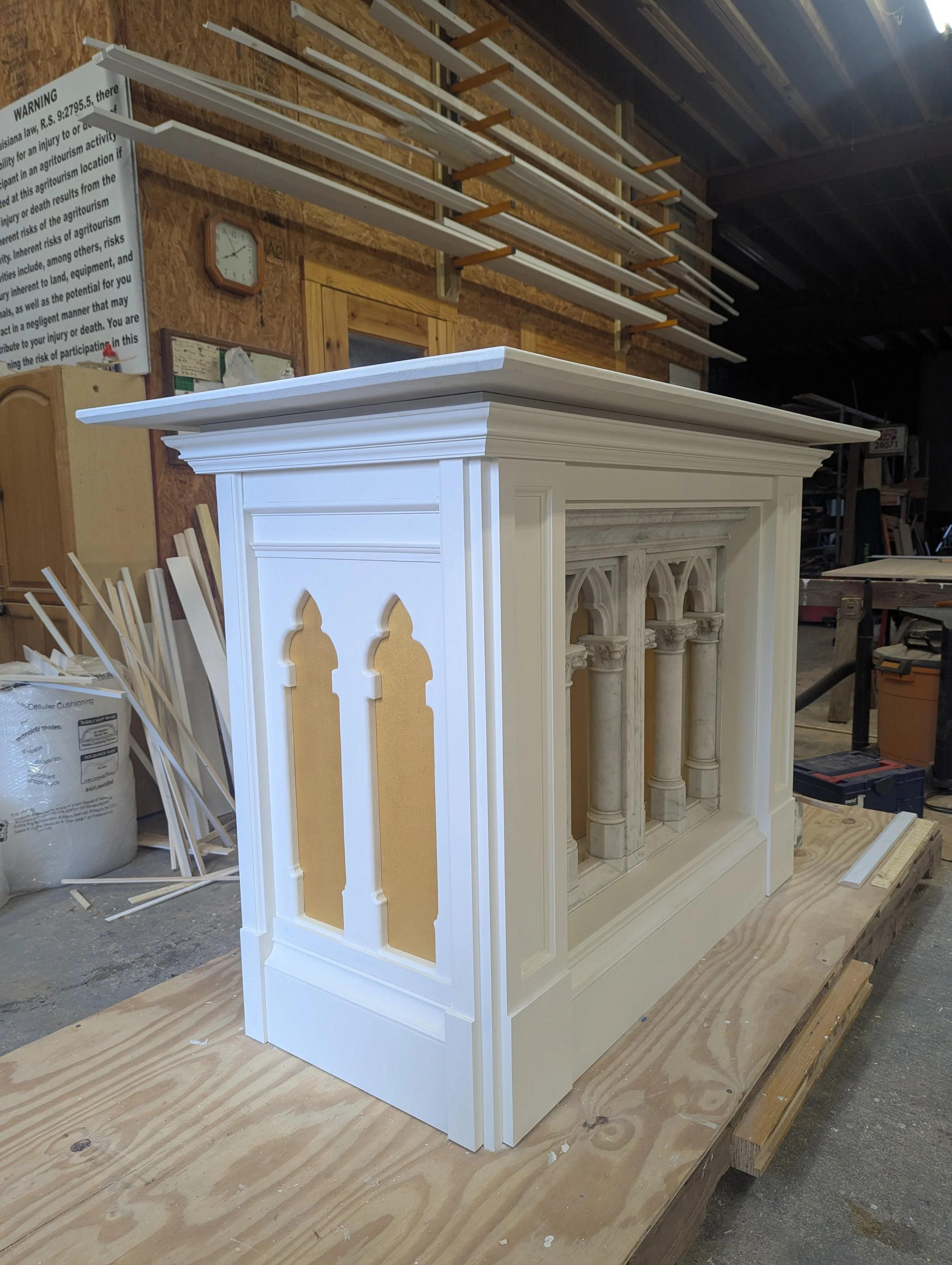A partially assembled white fireplace with decorative arches and columns, set on a wooden platform in a woodworking shop.