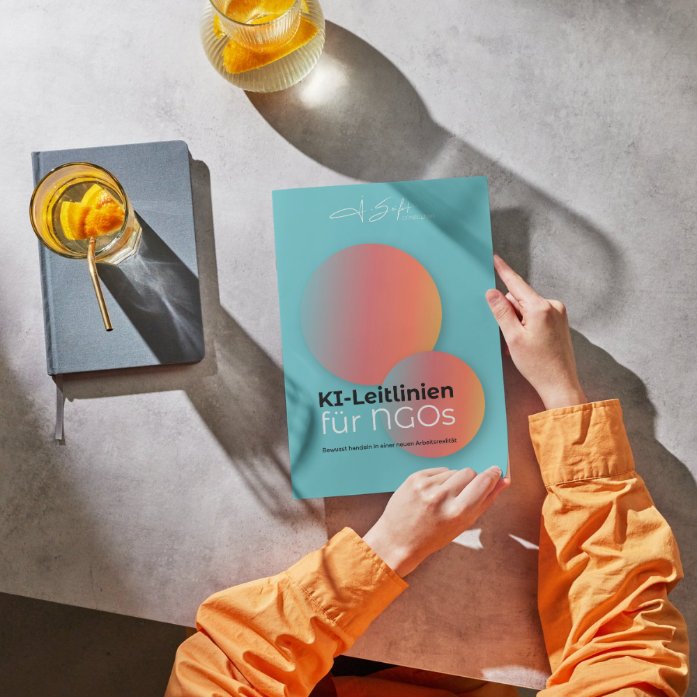 Eine Hand hält ein blaublaunes Heft mit dem Titel 'KI-Leitlinien für NGOs'. Auf dem Tisch vor der Person liegt ein grauer Notizblock mit einem Glas Wasser oder Saft mit Zitrusfrucht und ein weiteres Glas ebenfalls mit Zitrusfrucht. Im Hintergrund befindet sich eine weitere Zitrusfrucht in einem einfarbigen Gefäß.