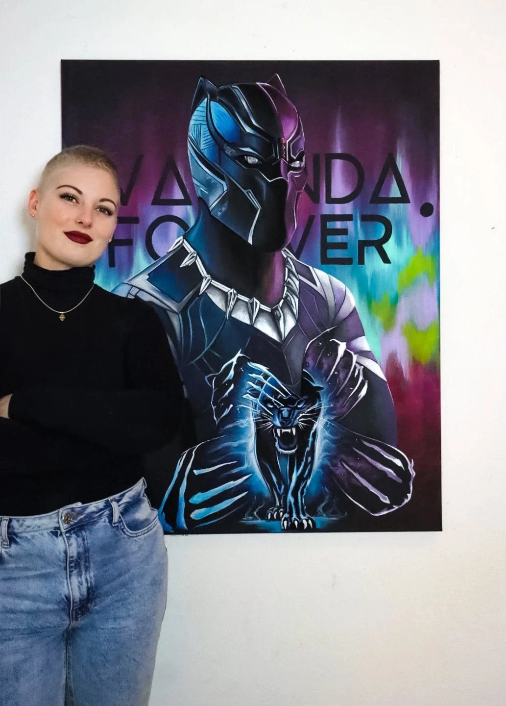 Eine junge Frau mit kurzen blonden Haaren, dunklem Lippenstift und schwarzen Kleidung steht neben einem schwarzen Panther Wandbild mit einem futuristischen schwarzen Panther-Krieger, der eine Halskette trägt, in einer Galerie.
