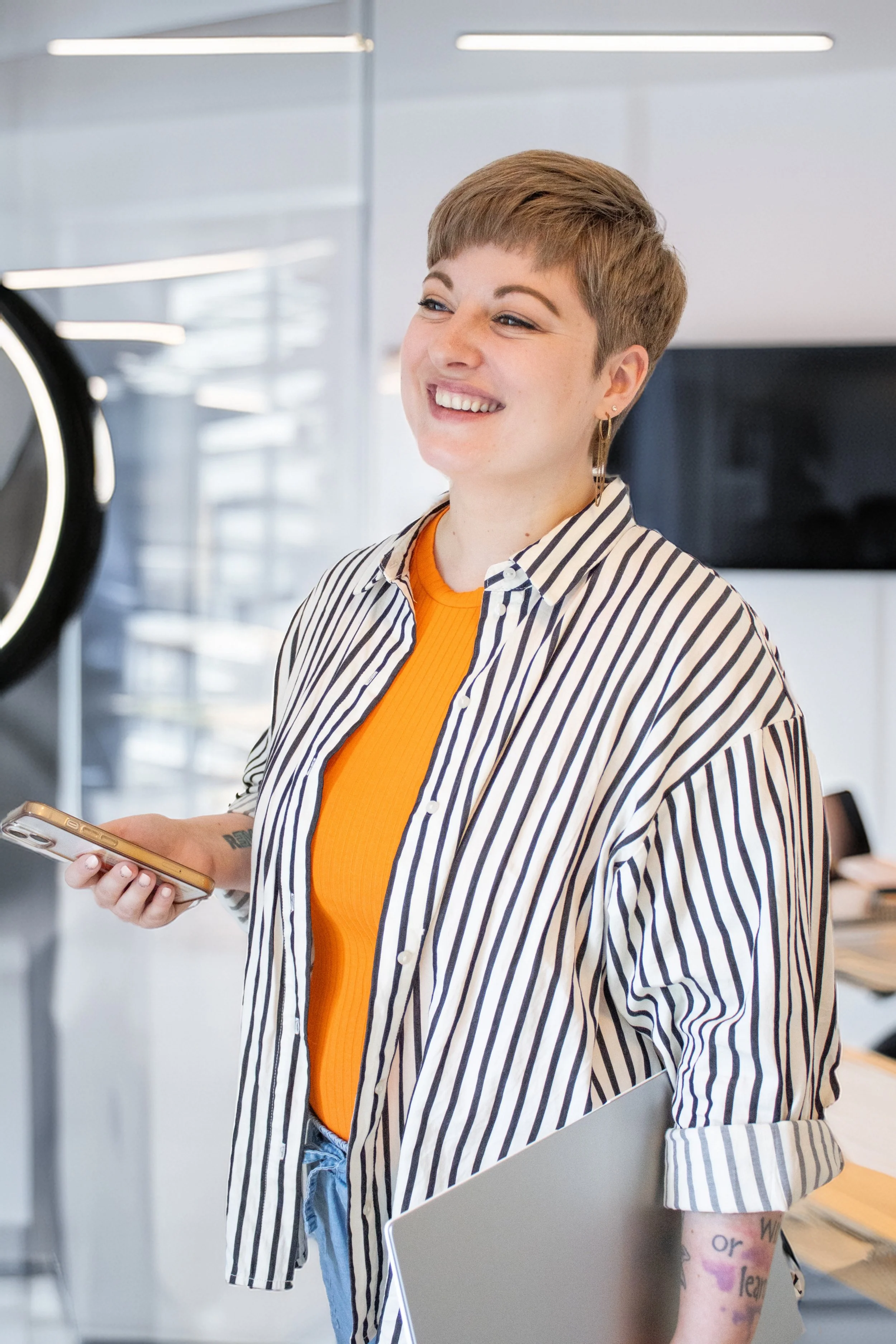 Junge Frau mit Kurzhaarschnitt, lächelnd, trägt eine gestreifte Bluse und ein orangefarbenes Shirt, hält ein Smartphone und einen Laptop in der Hand in einem modernen Büro.