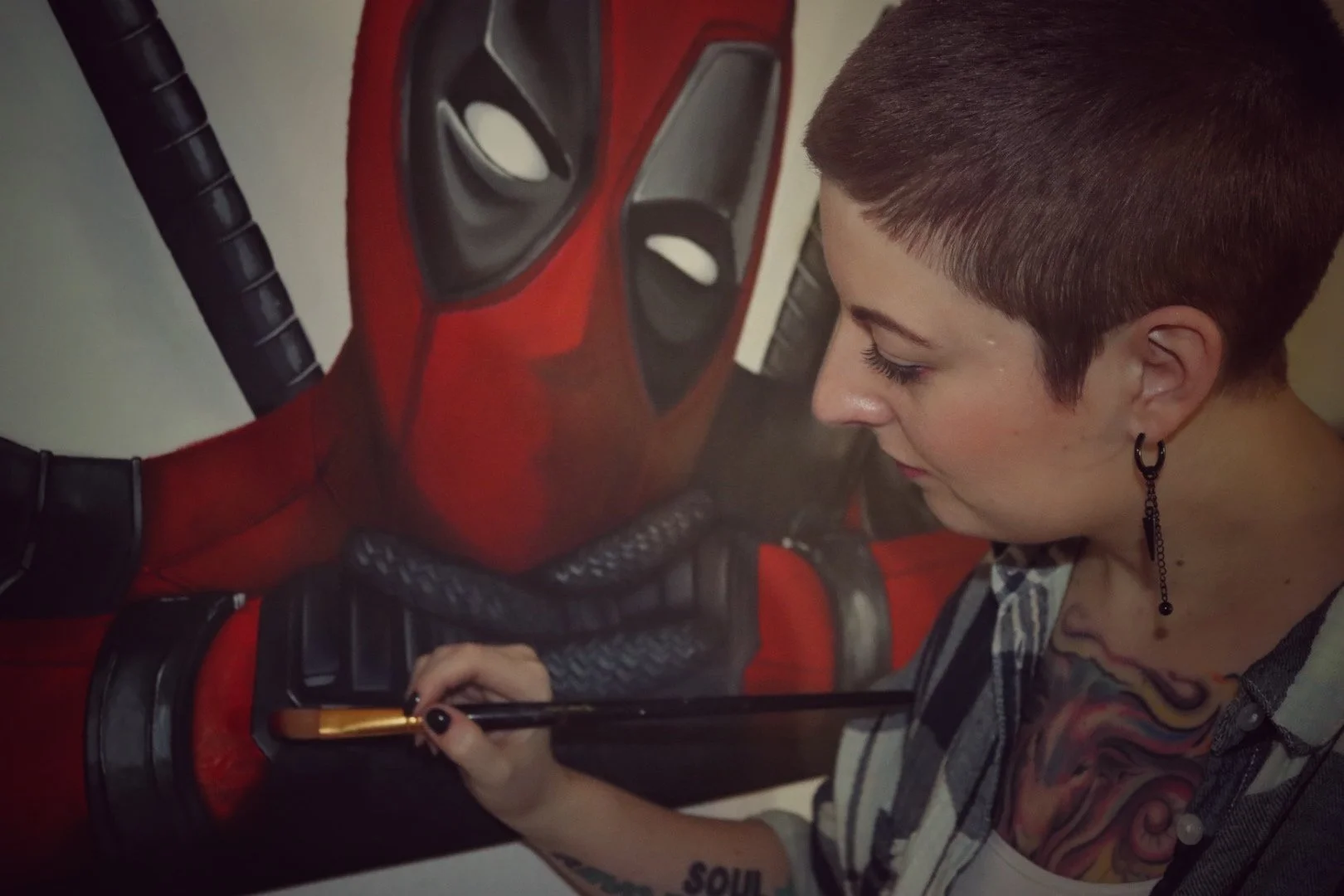 Künstlerin malt eine Wandzeichnung von Deadpool, eine Comic-Charakter, mit Pinsel. Die Künstlerin hat kurze braune Haare, trägt ein kariertes Hemd, einen weißen Untergrund und eine auffällige Tätowierung am Hals.