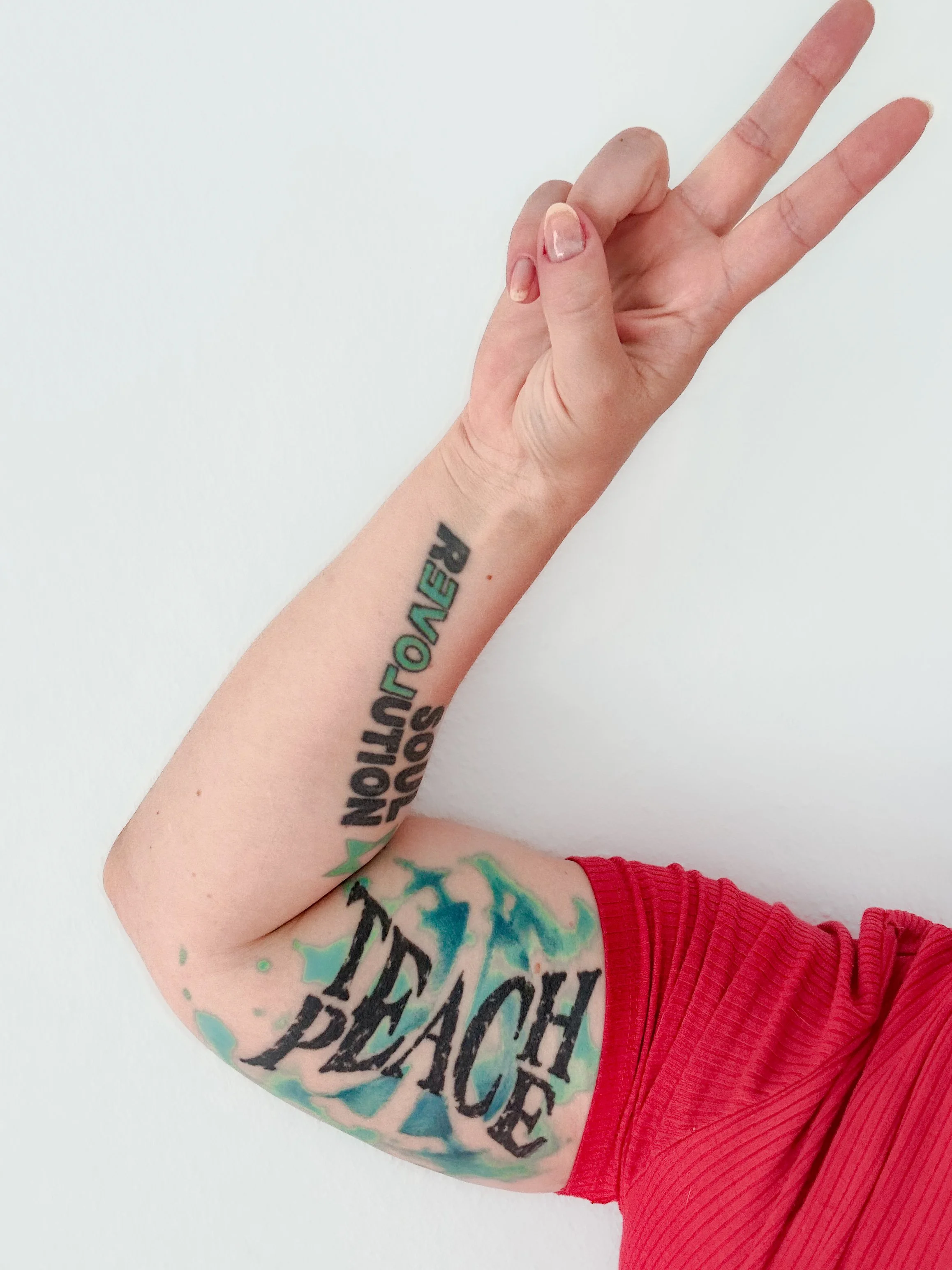 Ein Arm mit Tattoos, der eine Peace-Geste macht, trägt ein rotes Shirt gegen eine weiße Wand.