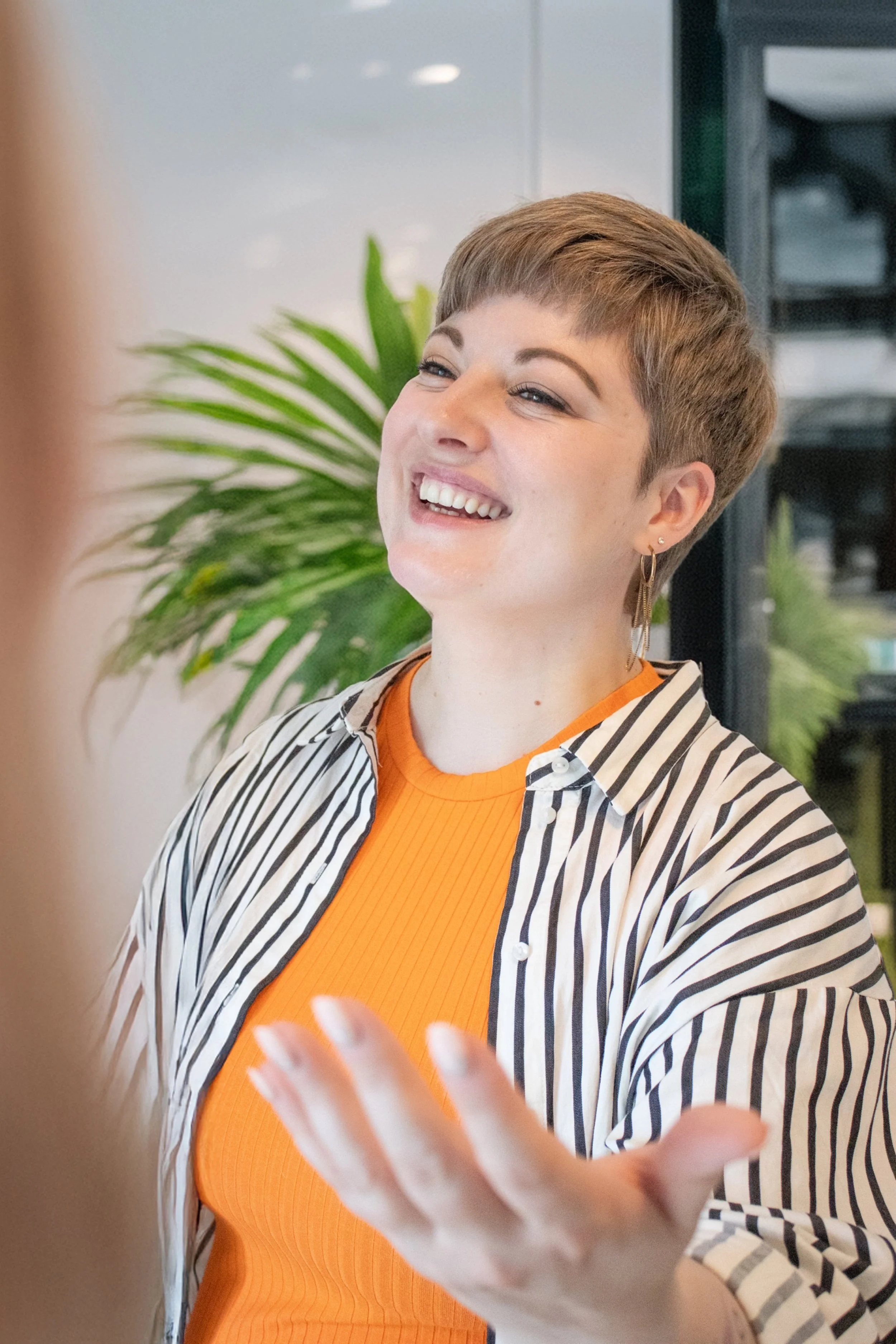 Eine lachende Frau mit kurzen braunen Haaren, die ein orangefarbenes Oberteil und eine schwarz-weiße gestreifte Jacke trägt, in einem modernen, hell erleuchteten Raum mit Pflanzen im Hintergrund.